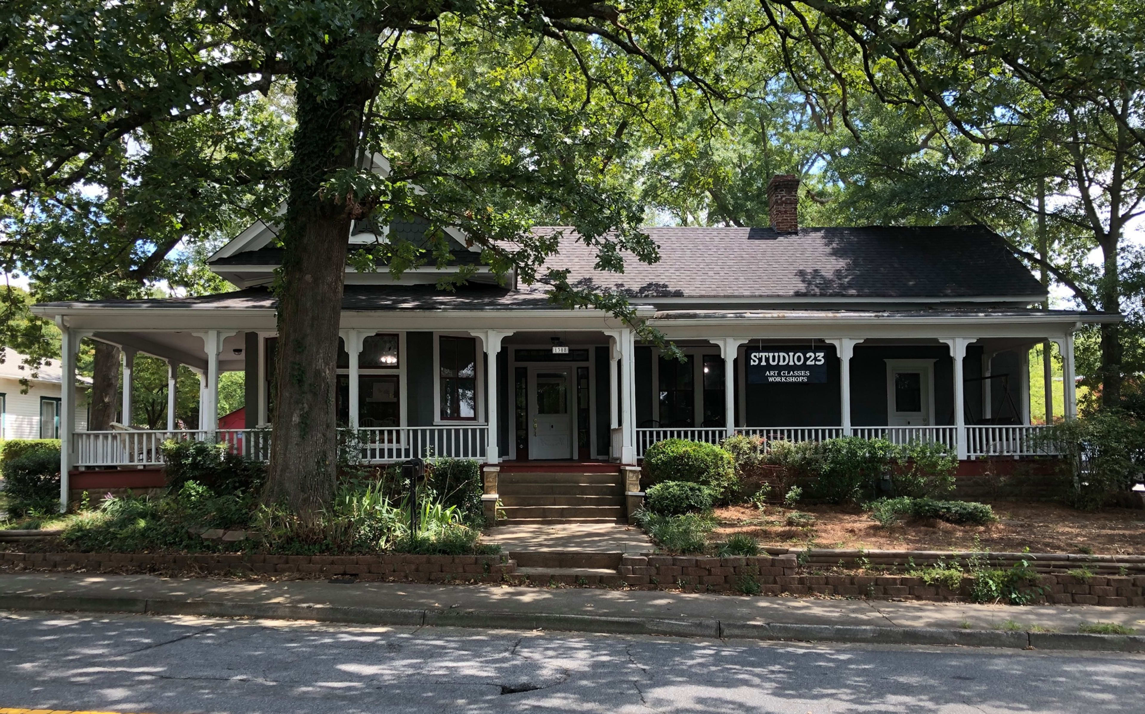 The image shows a large, one-story house with a prominent front porch, surrounded by trees and greenery, and featuring a sign that reads "STUDIO 23."