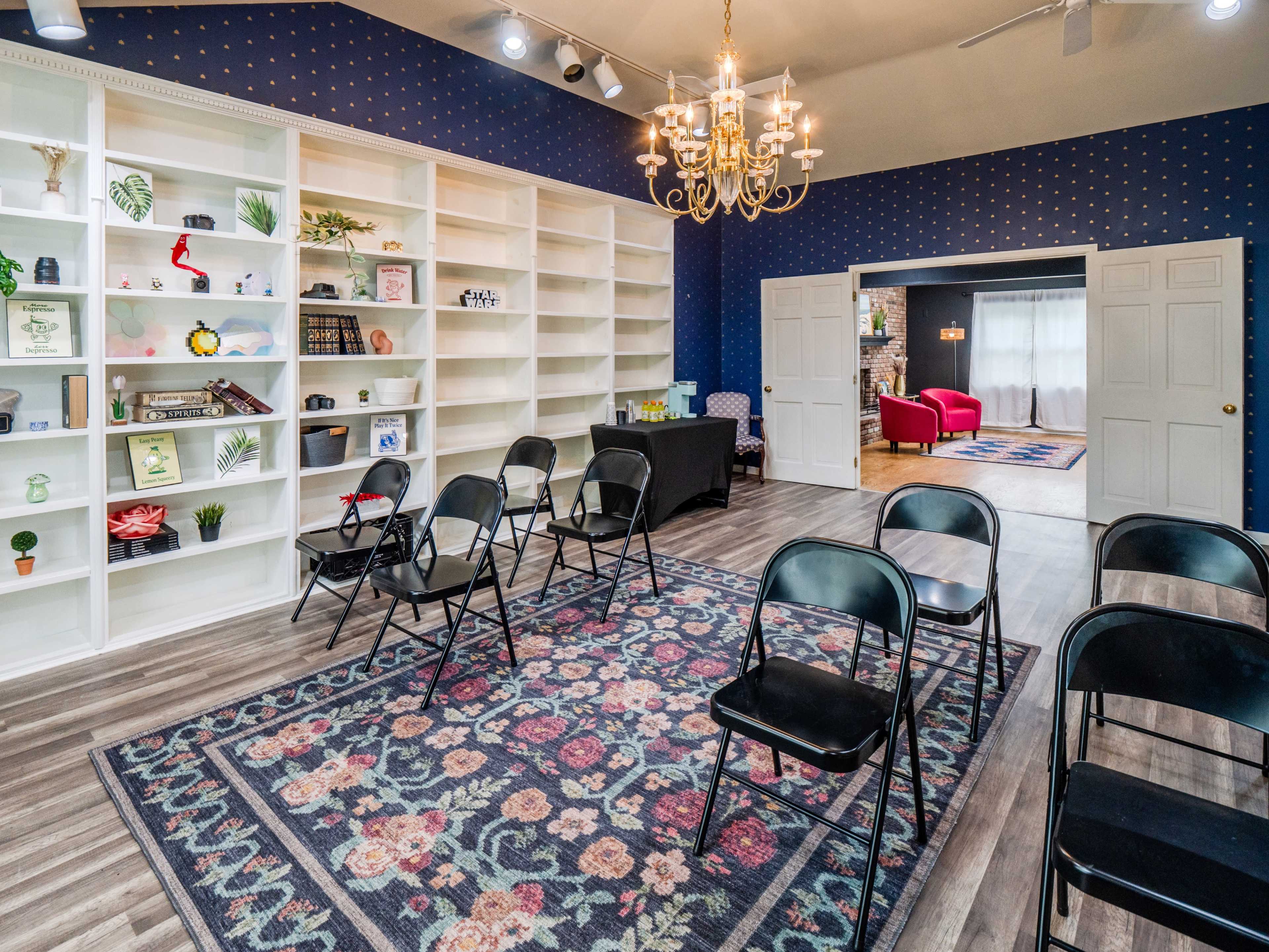 A room featuring a large white bookshelf filled with various items, a central area with black chairs arranged around a patterned rug, and a doorway leading to a separate, brightly lit room with a red chair.