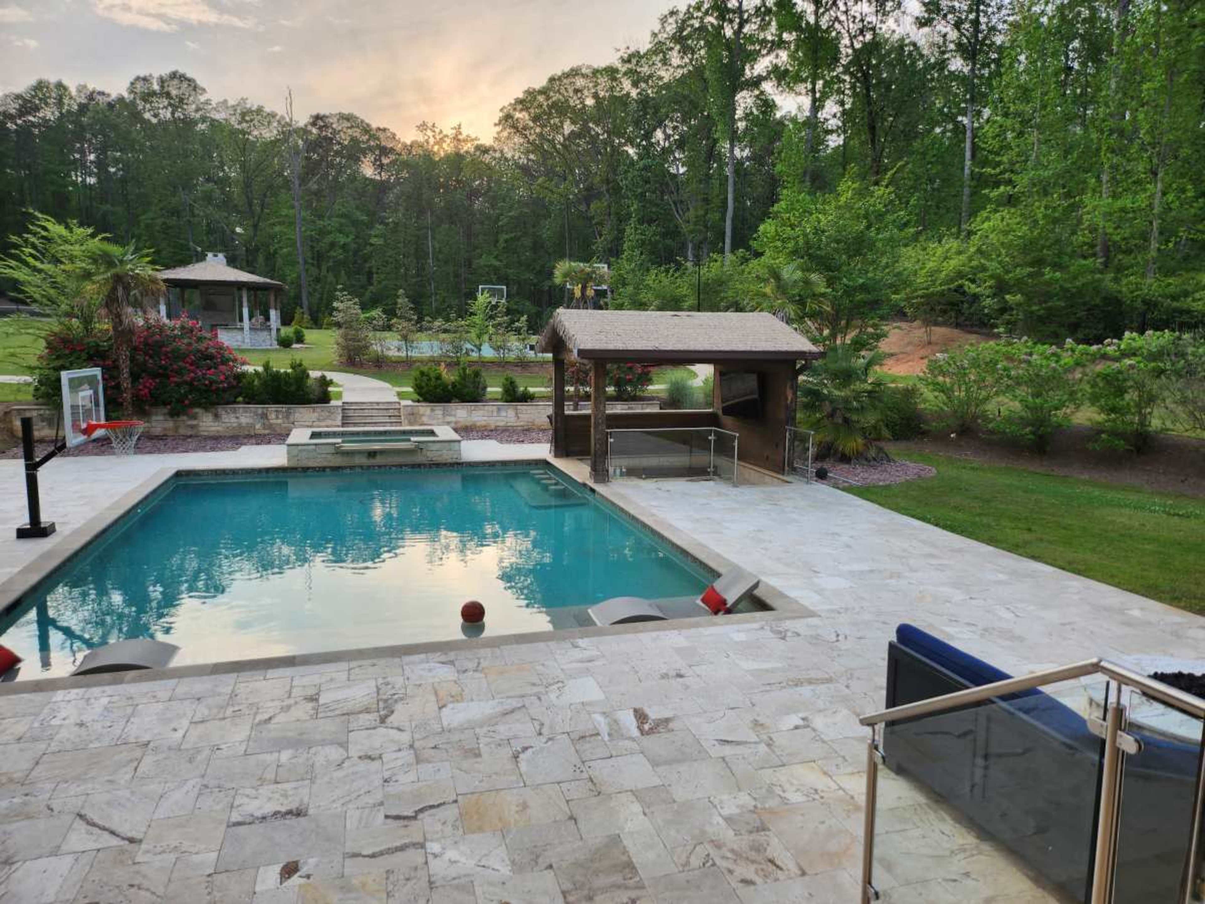 A rectangular swimming pool surrounded by a stone patio, with a covered seating area and lush greenery in the background.
