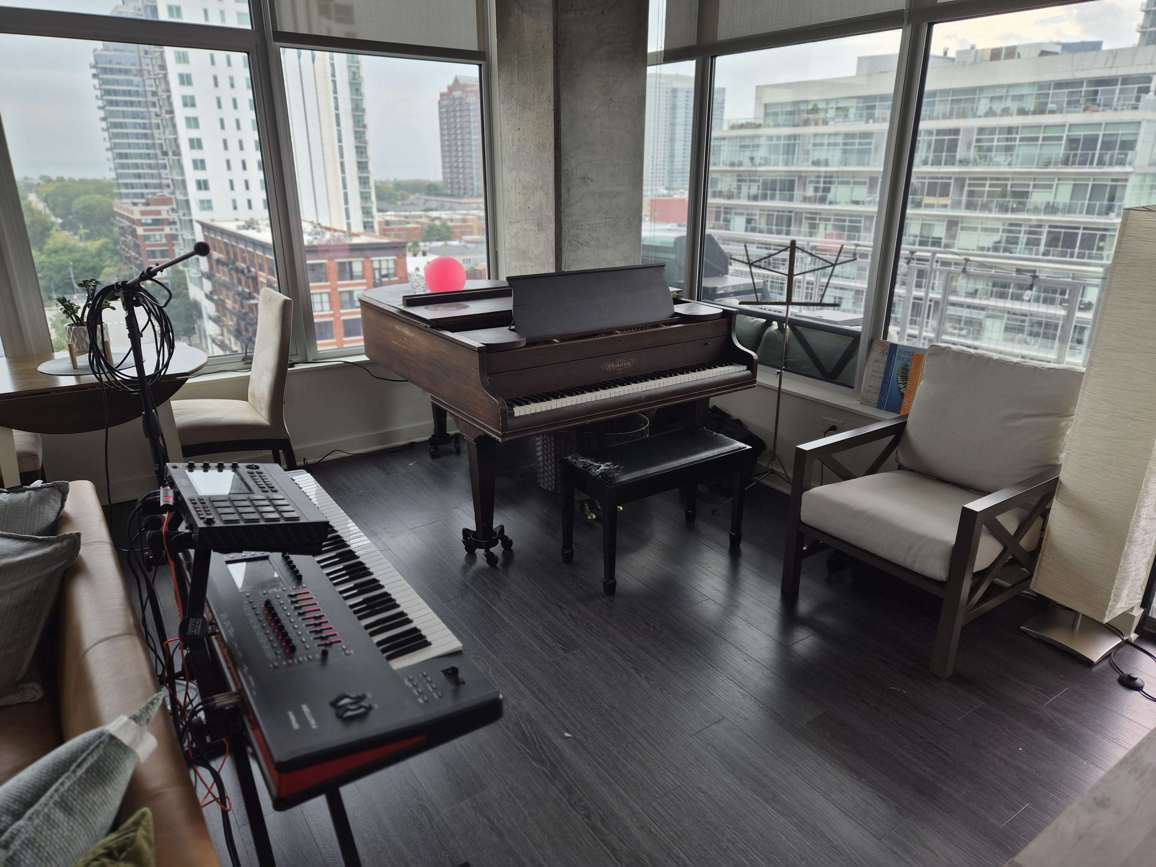 The scene features a music corner in a modern apartment with a grand piano, a keyboard, and comfortable seating, surrounded by large windows showcasing a city view.