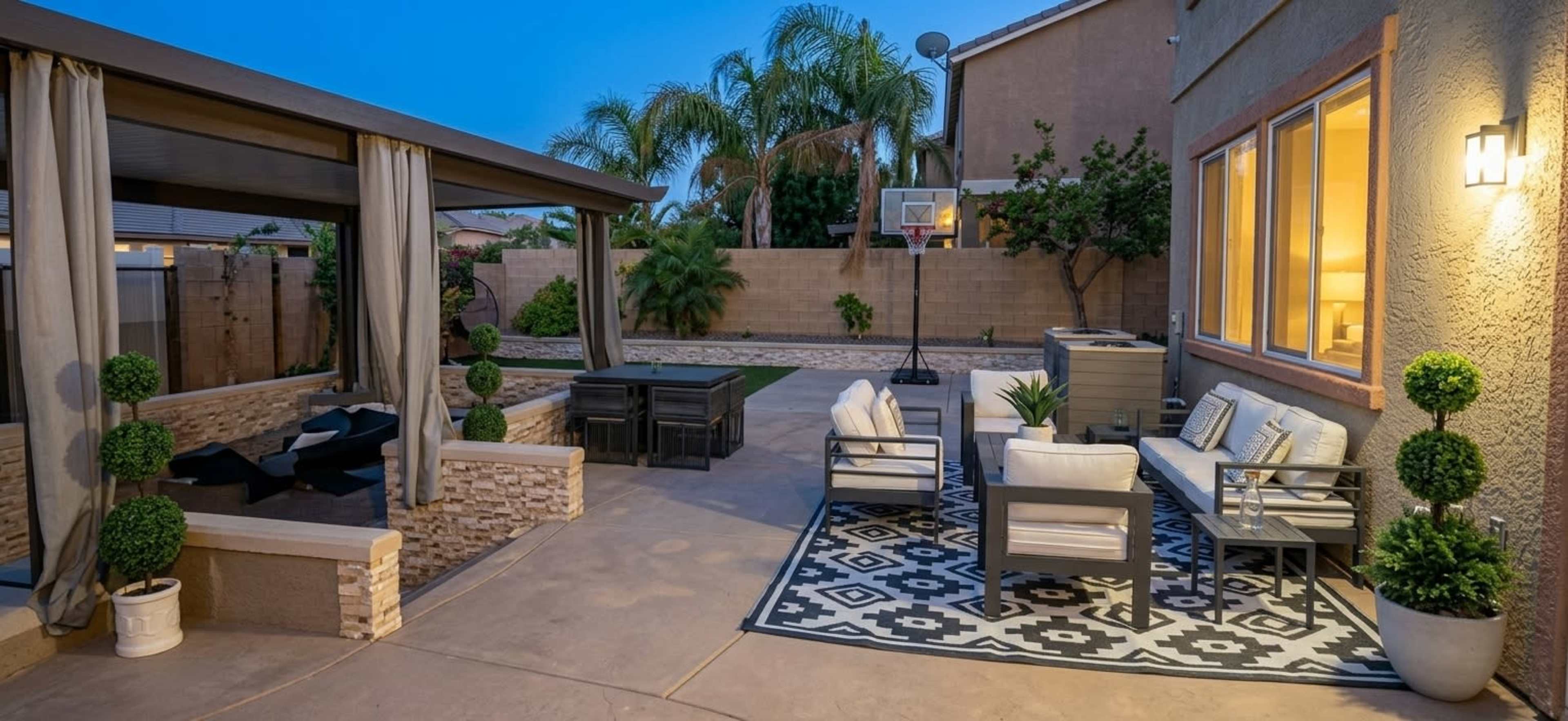 The image shows a backyard patio area with seating furniture, a decorative rug, a basketball hoop, and landscaped greenery.