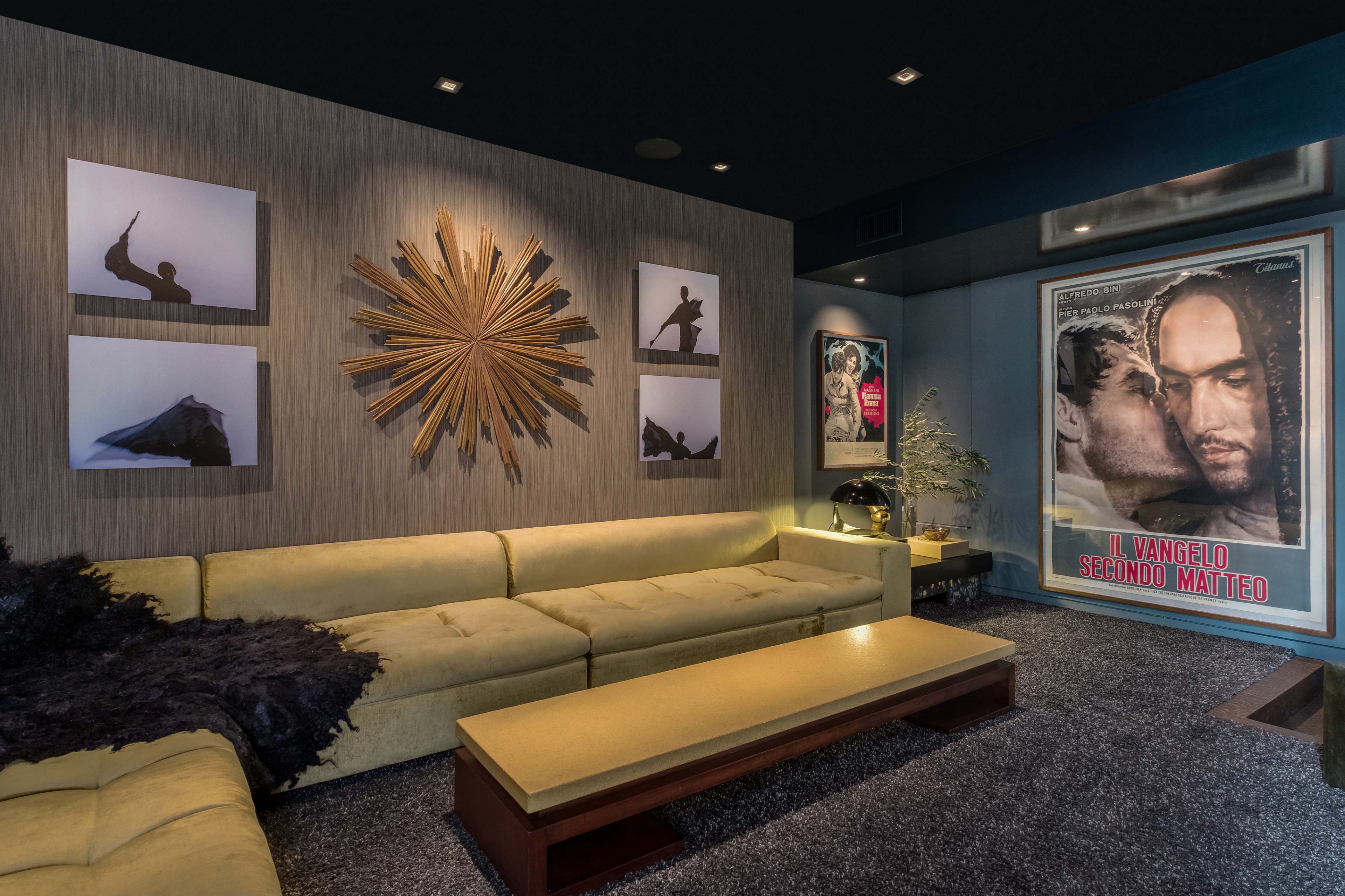 The image shows a modern living room featuring a yellow sectional couch, a coffee table, framed artwork on the walls, and a large movie poster.