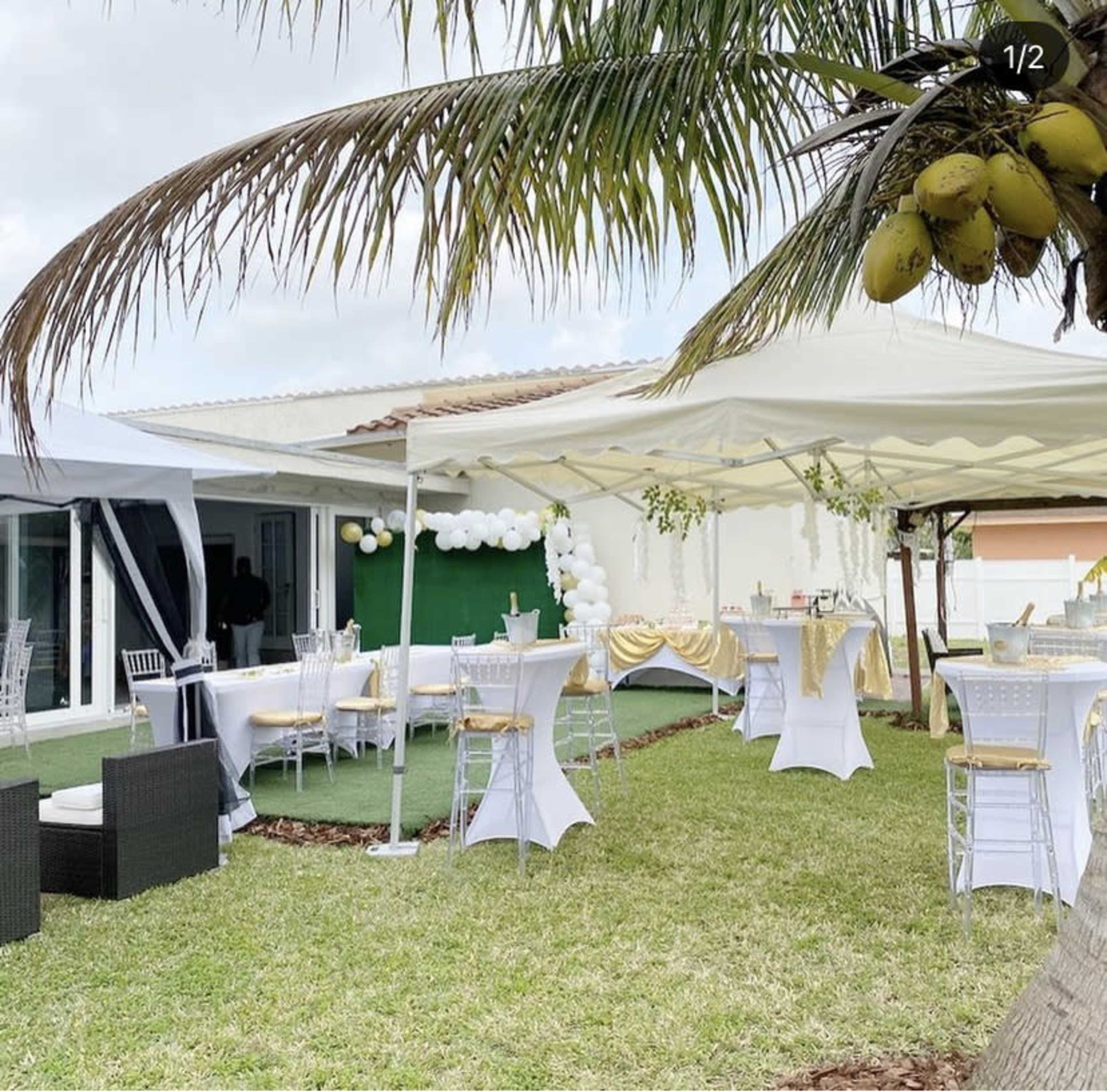 The scene shows a outdoor event space with white tables and chairs set up under canopies, surrounded by grass and palm trees.
