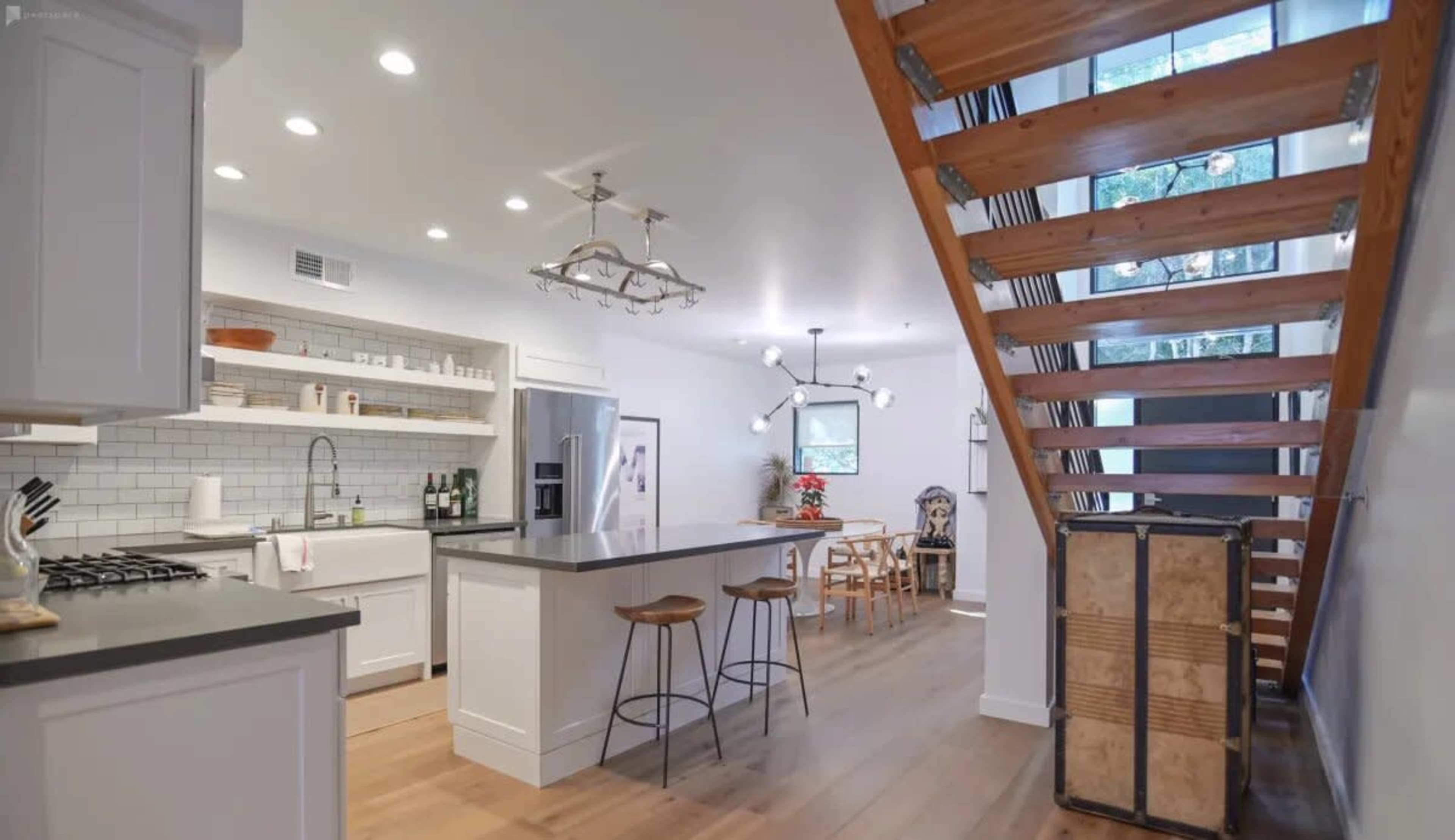 A modern kitchen features white cabinetry and an island with bar stools, adjacent to a wooden staircase leading to an upper floor.