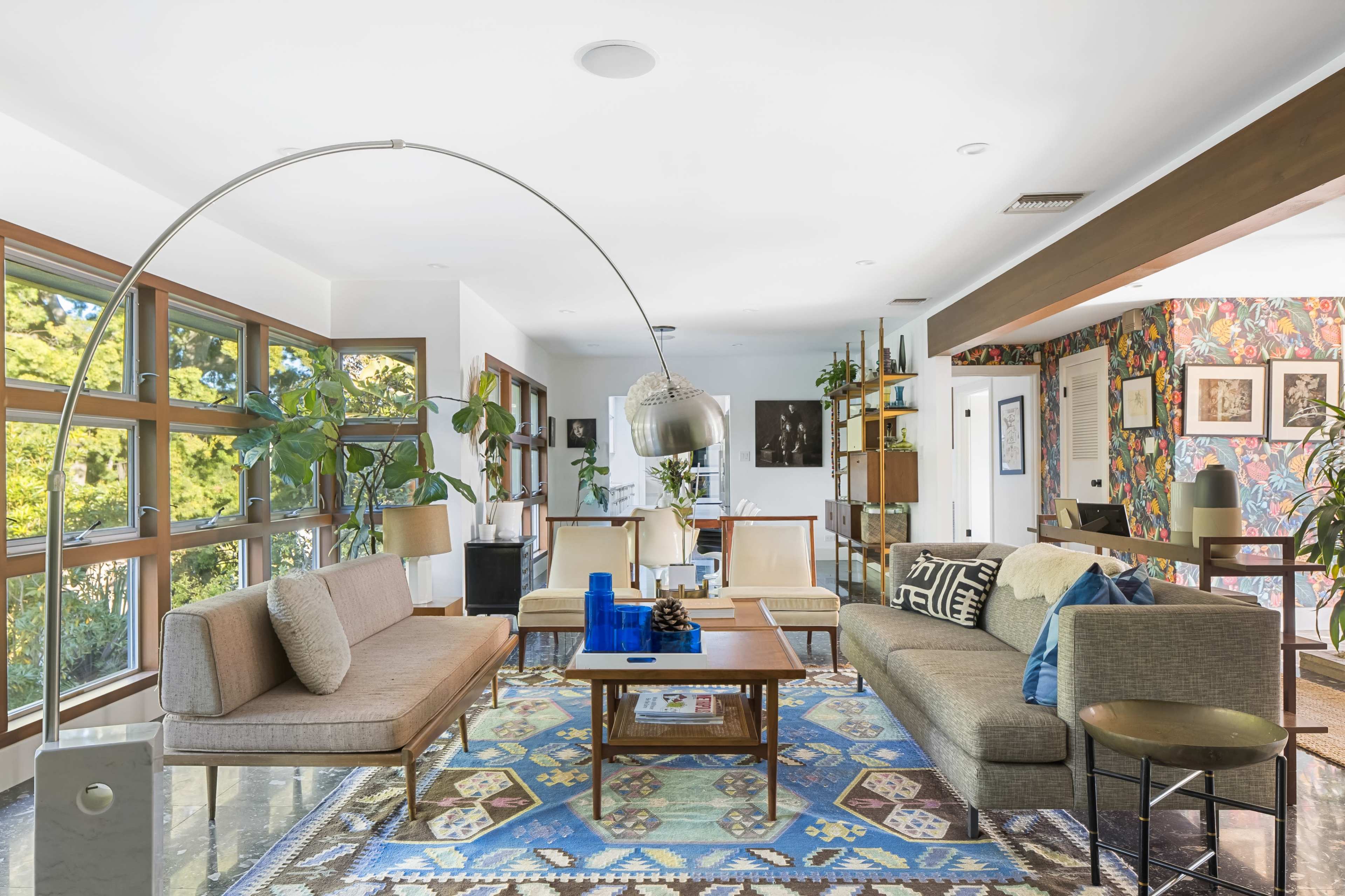 A modern living room features large windows, a curved floor lamp, and a colorful patterned rug beneath a coffee table surrounded by various seating options.