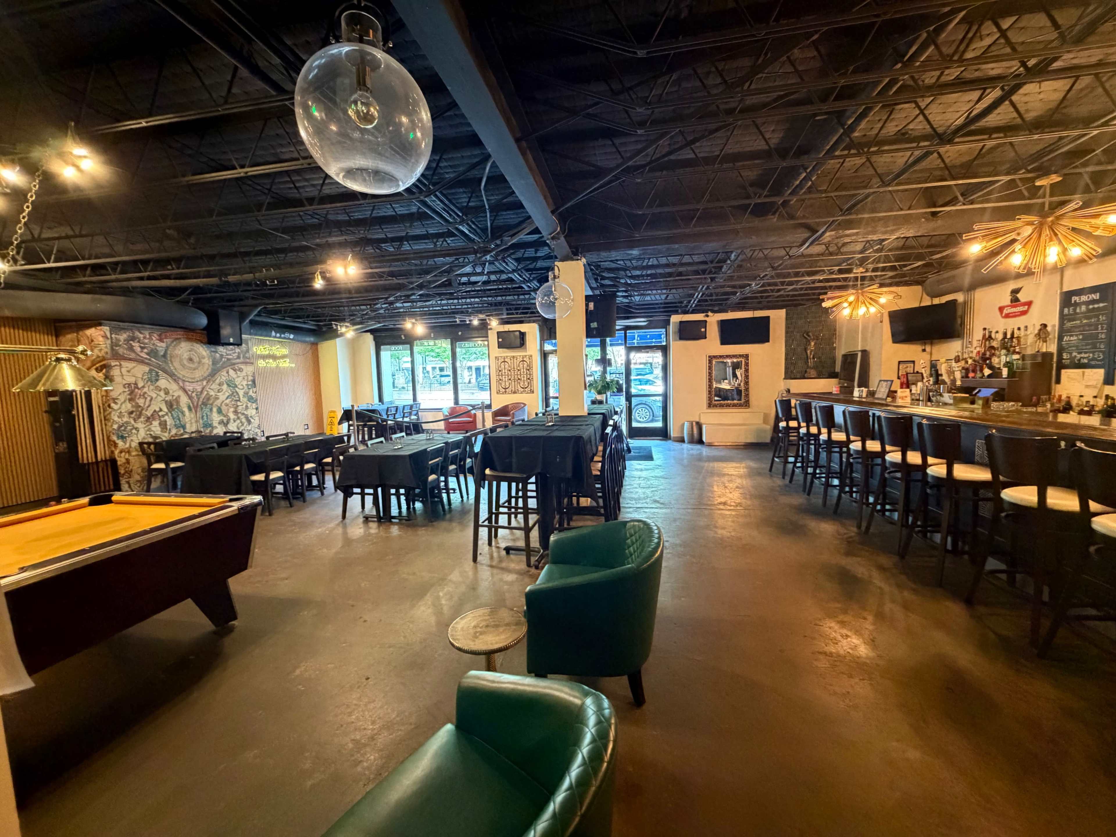 The interior of a spacious, modern bar features a pool table, a seating area with green chairs, and a long bar counter.
