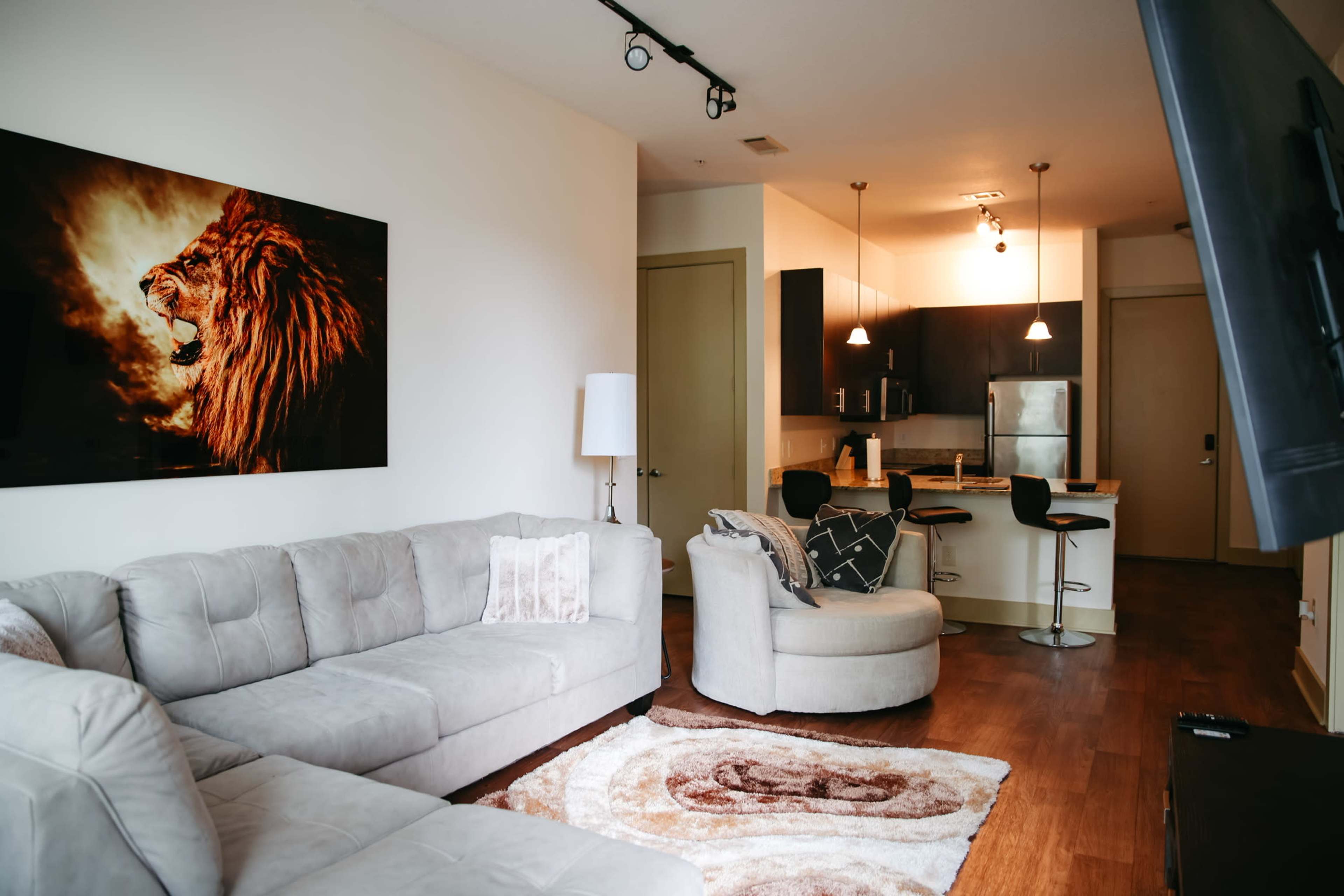 The image shows a modern living room with a grey sectional sofa, a round chair, a large lion portrait on the wall, and an open kitchen area in the background.