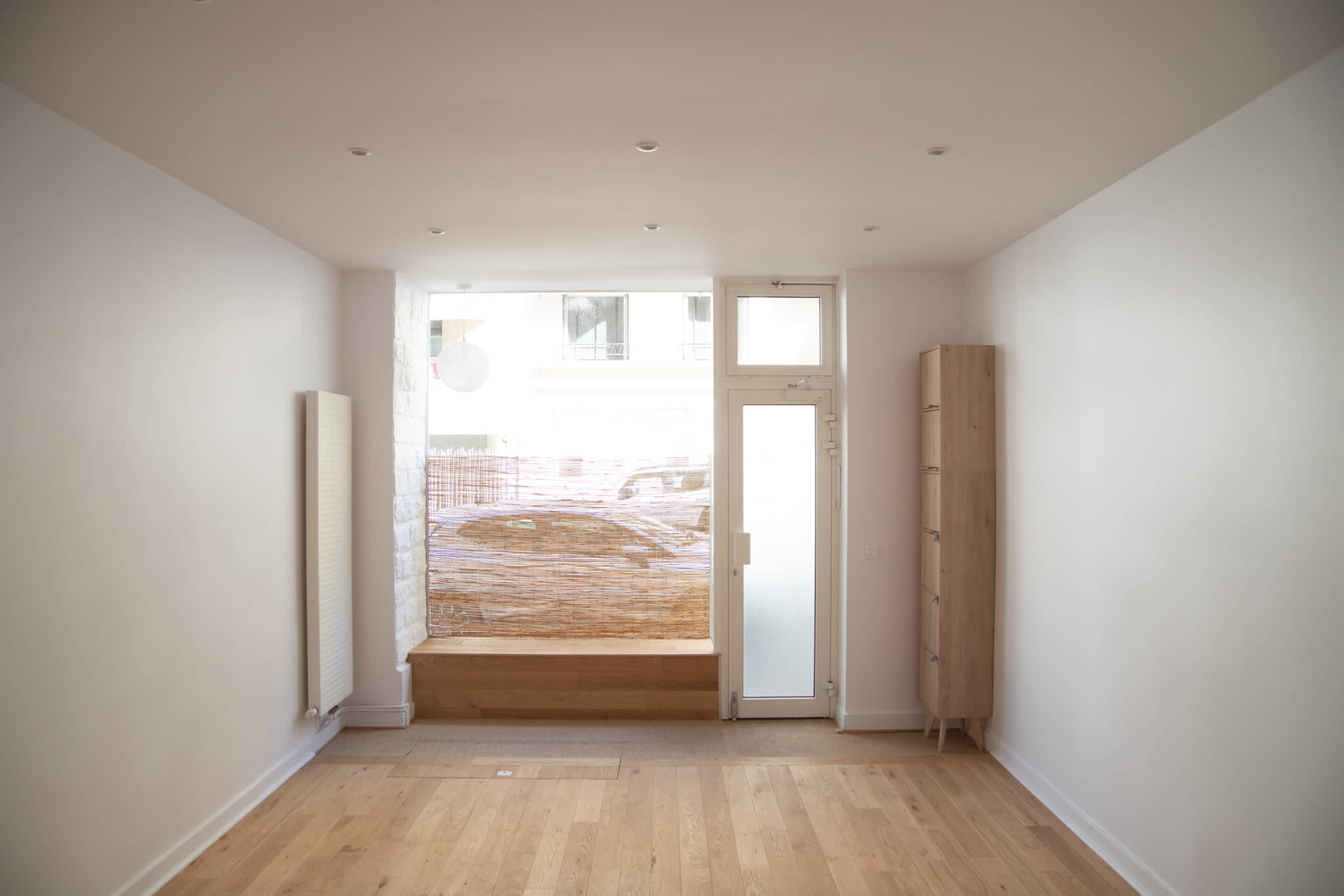 A minimalist room with wooden flooring, a large window, and a wooden shelf against a white wall.