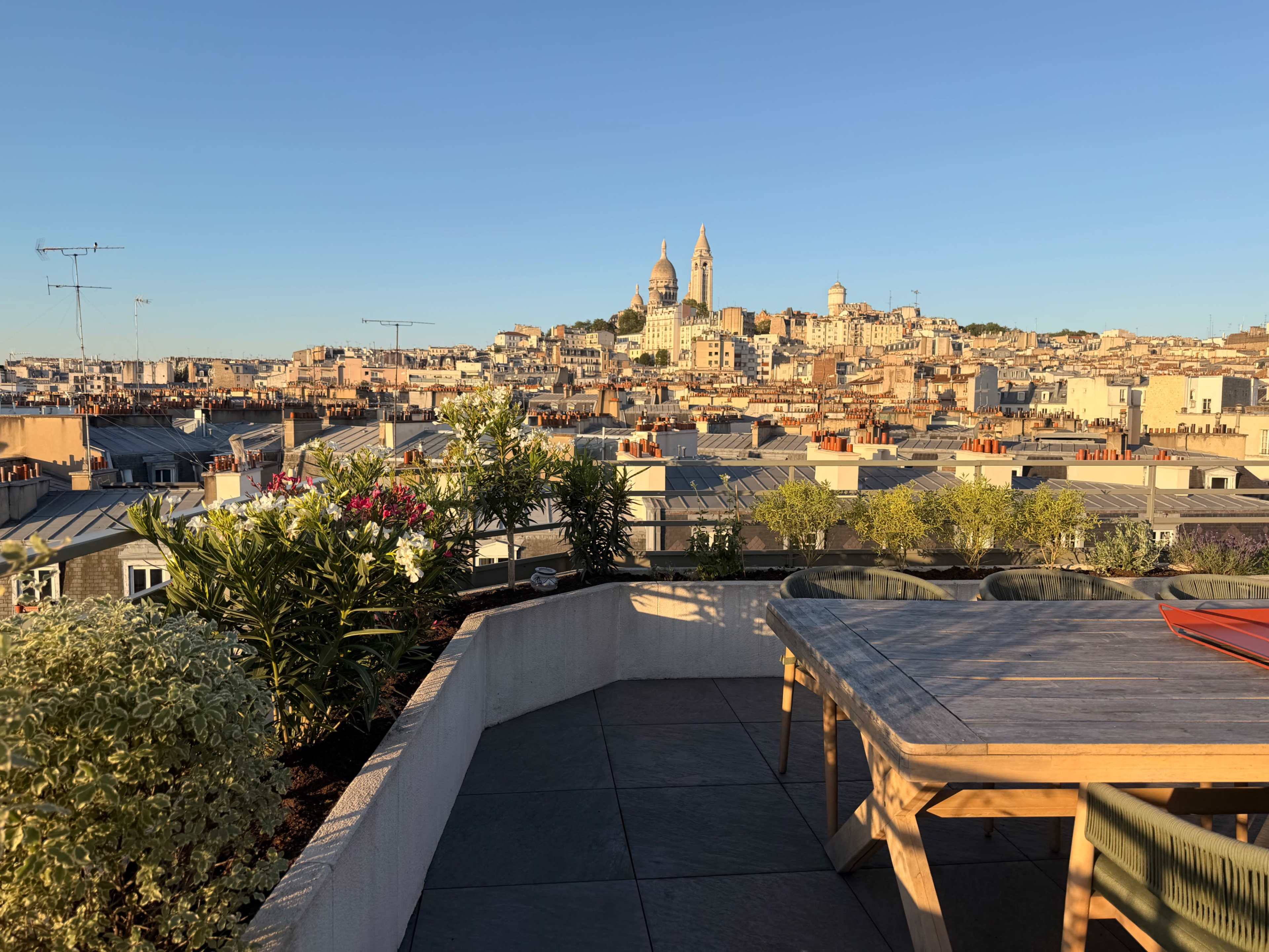 The image shows a rooftop terrace with a table overlooking a cityscape featuring buildings and a distant church with towers in clear sky.