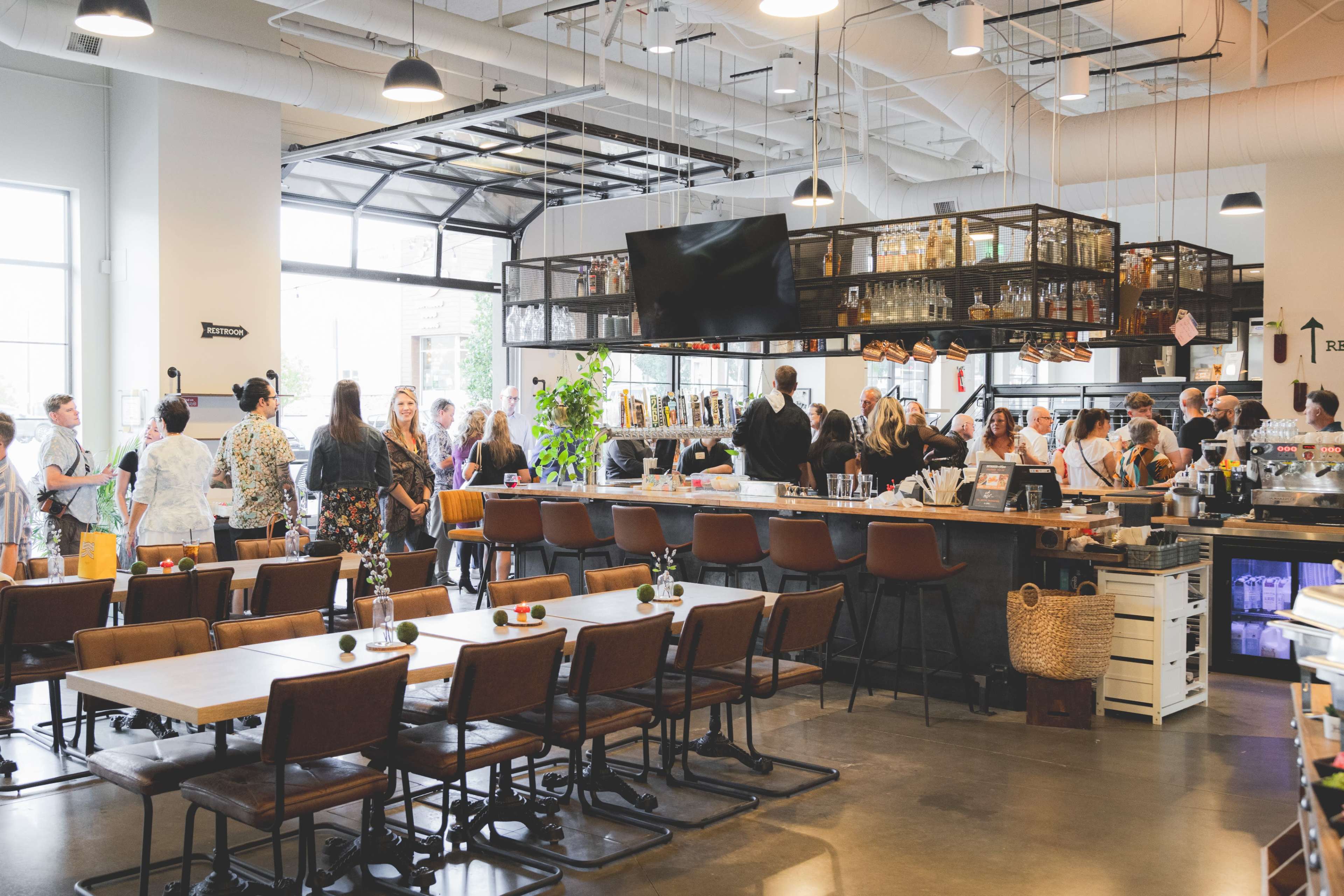 The image shows a bustling café interior with patrons seated at tables and others gathered around the bar area, featuring a variety of beverages and décor.