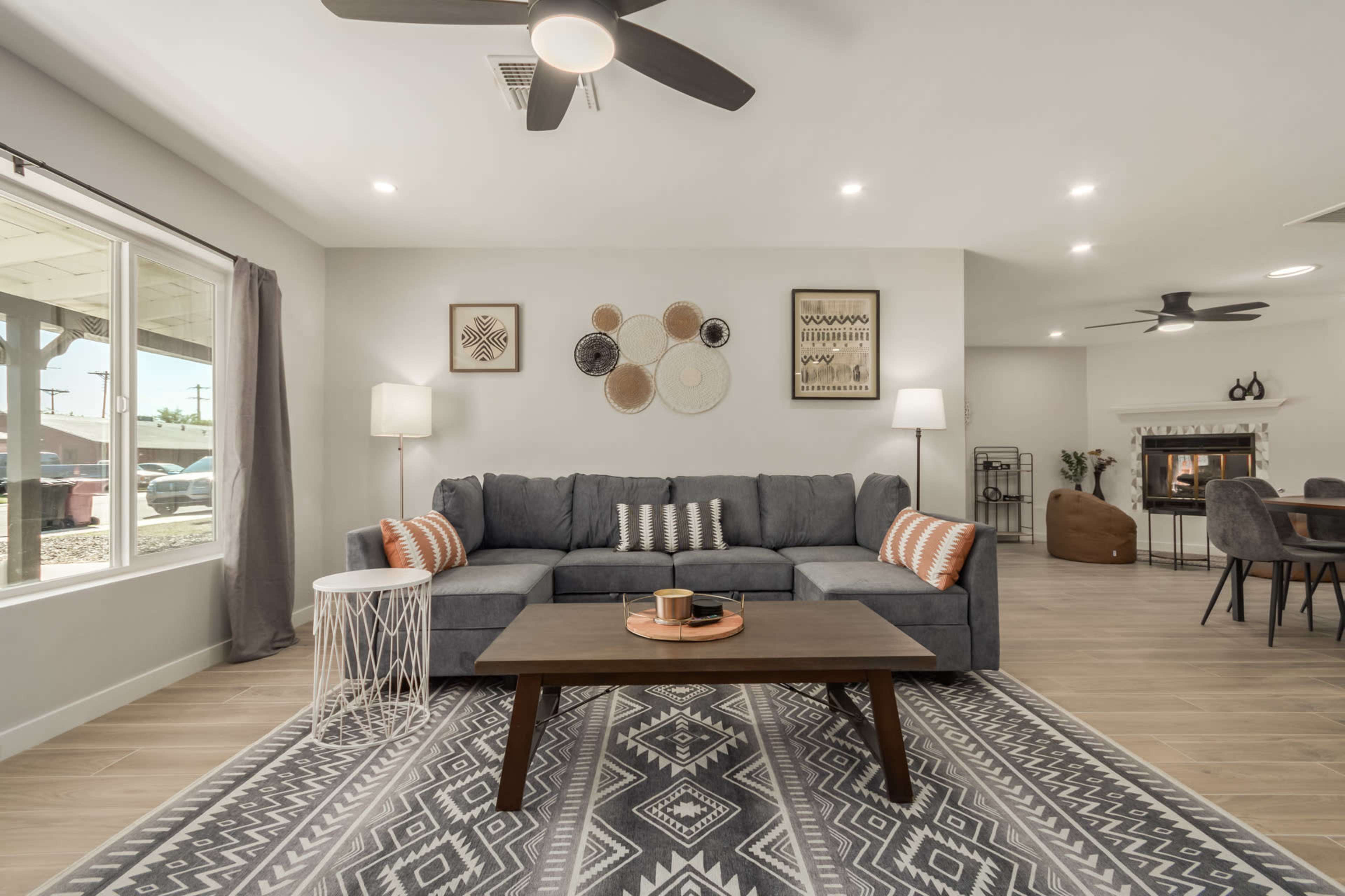 A modern living room features a grey sectional sofa, a wooden coffee table, decorative wall hangings, and large windows allowing natural light.