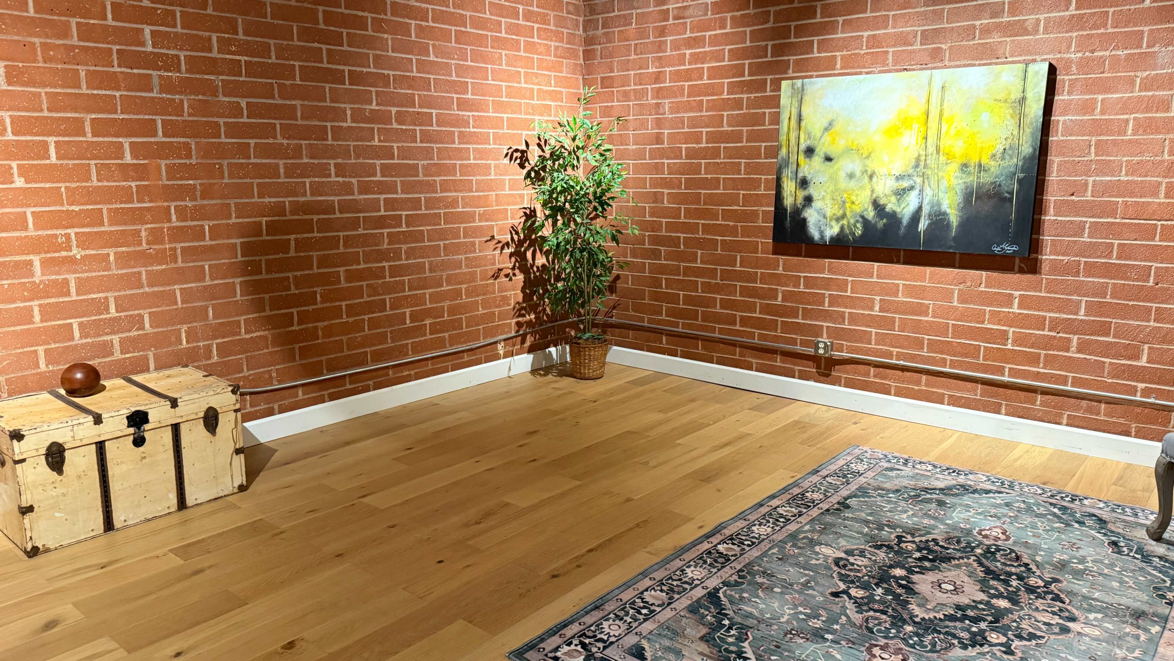 A room features a brick wall, a large painting with yellow and dark hues, a potted plant, a wooden trunk, and an area rug on a hardwood floor.