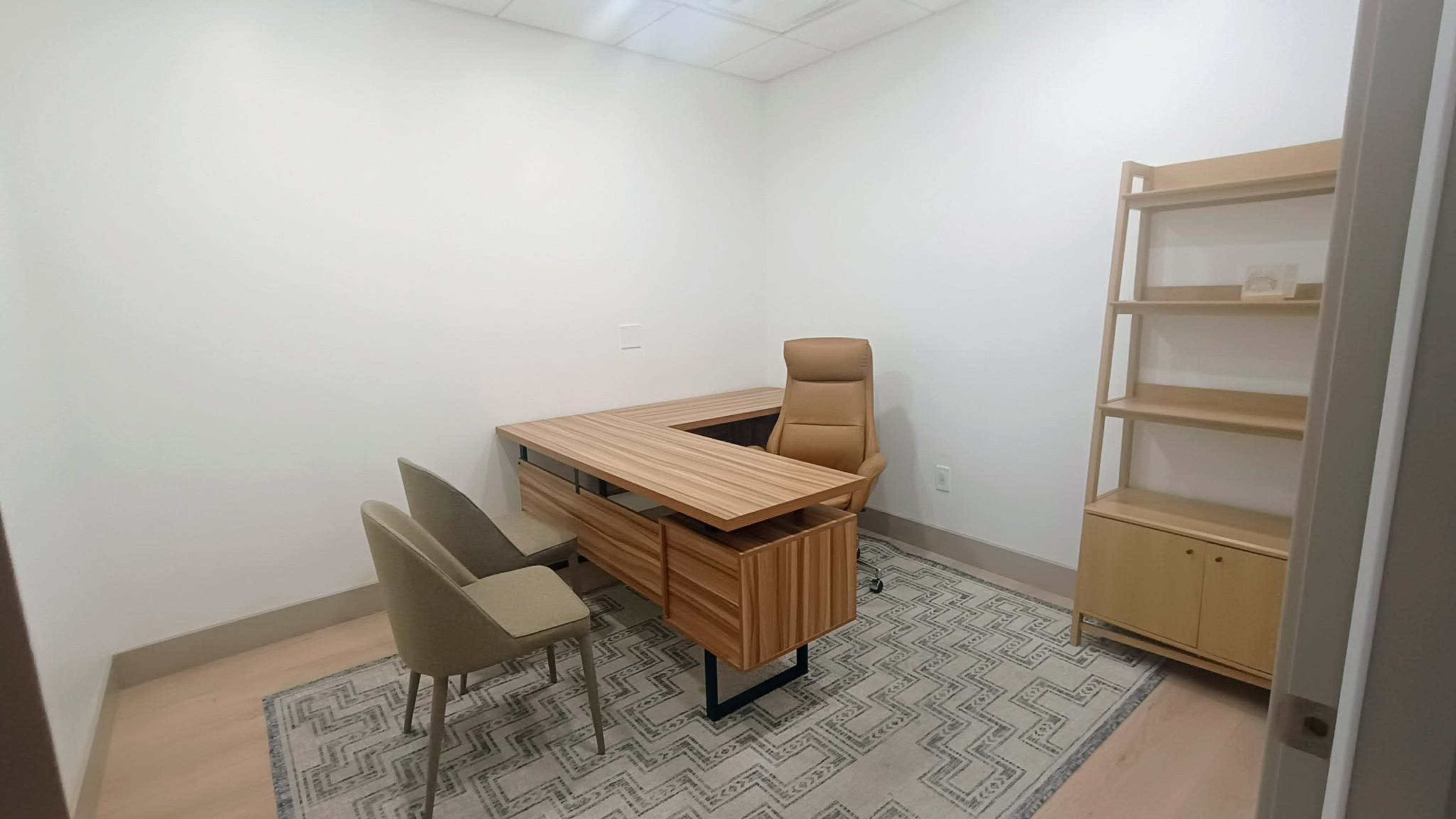 The image shows a minimalist office space with a wooden desk, a reclining chair, two beige chairs, and a shelf against a white wall.