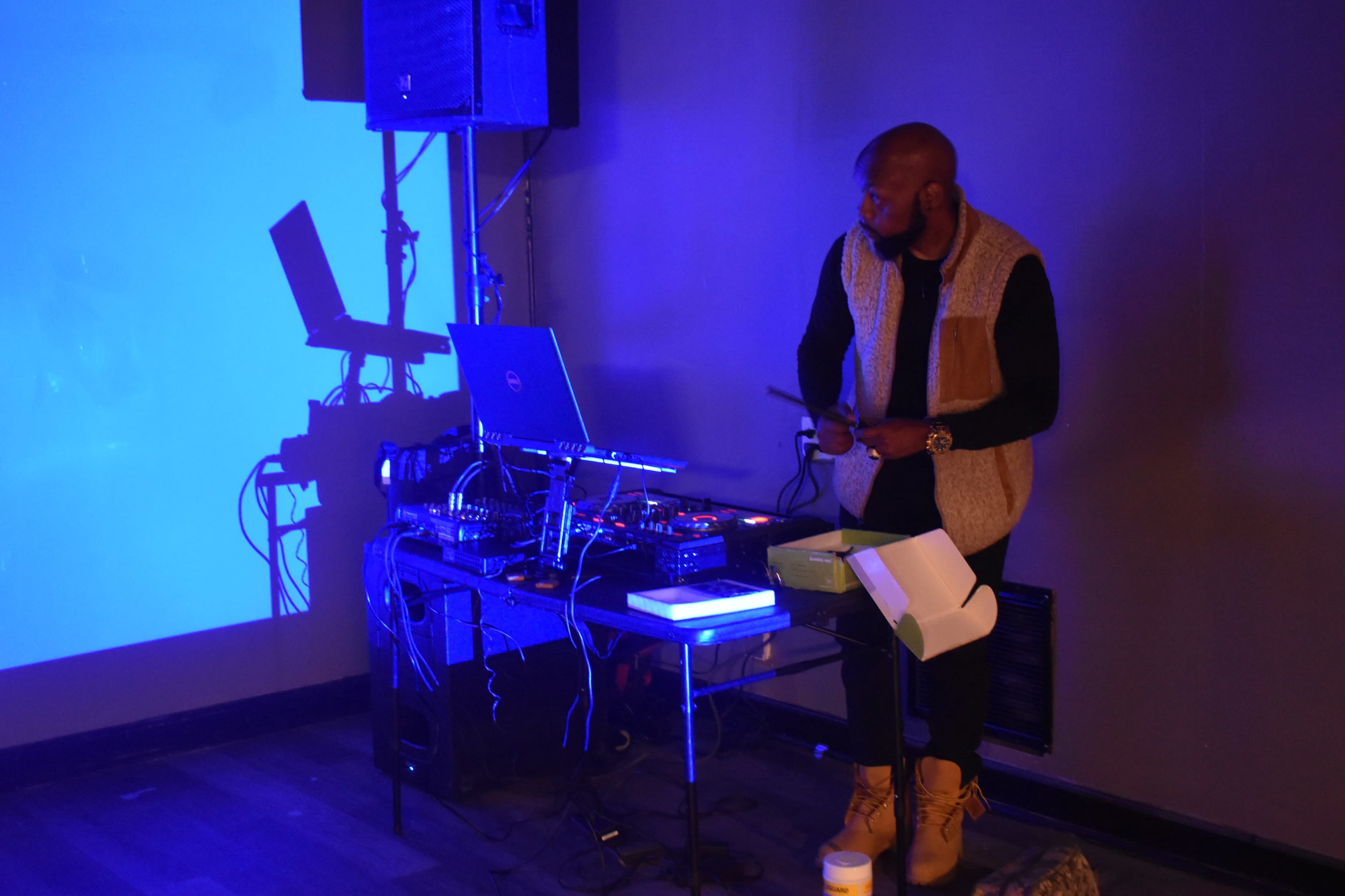 A DJ stands behind a table with turntables and a laptop, set against a wall illuminated by blue light.