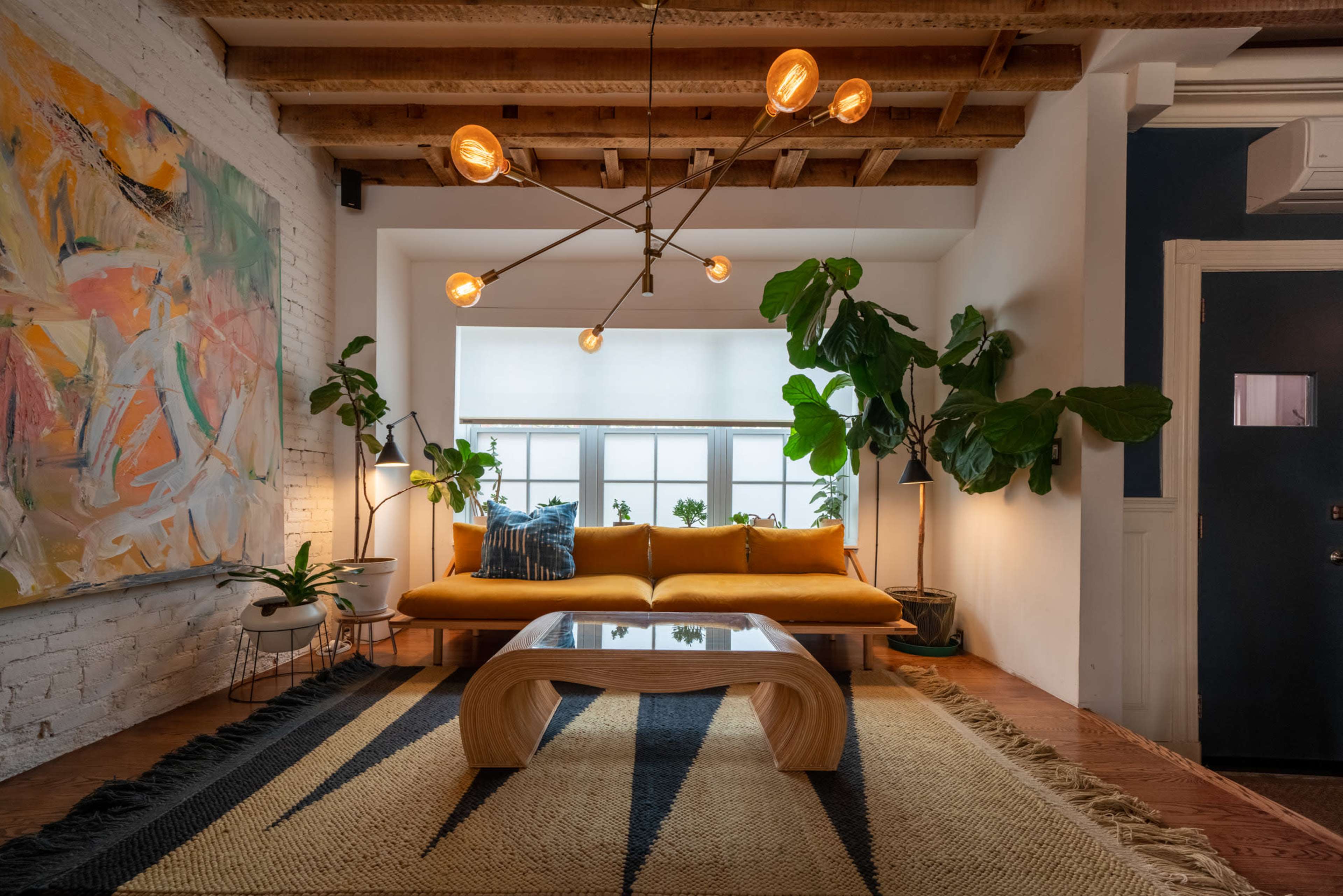 A modern living room featuring a yellow sofa, a geometric wooden coffee table, plants, and artwork on the walls under a wooden beam ceiling.