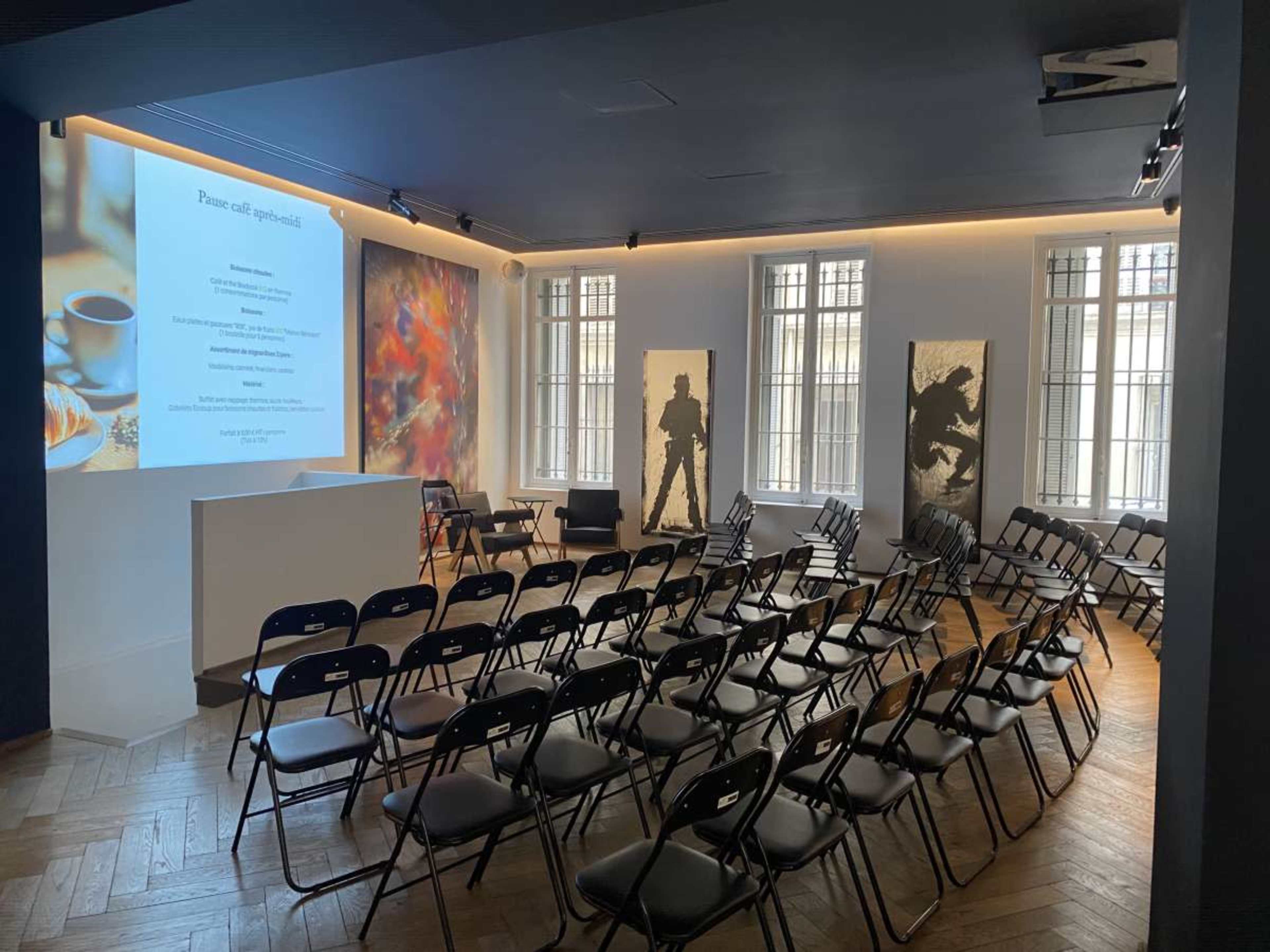 The image shows an indoor event space set up with rows of black chairs facing a presentation screen, flanked by large artworks on the walls.