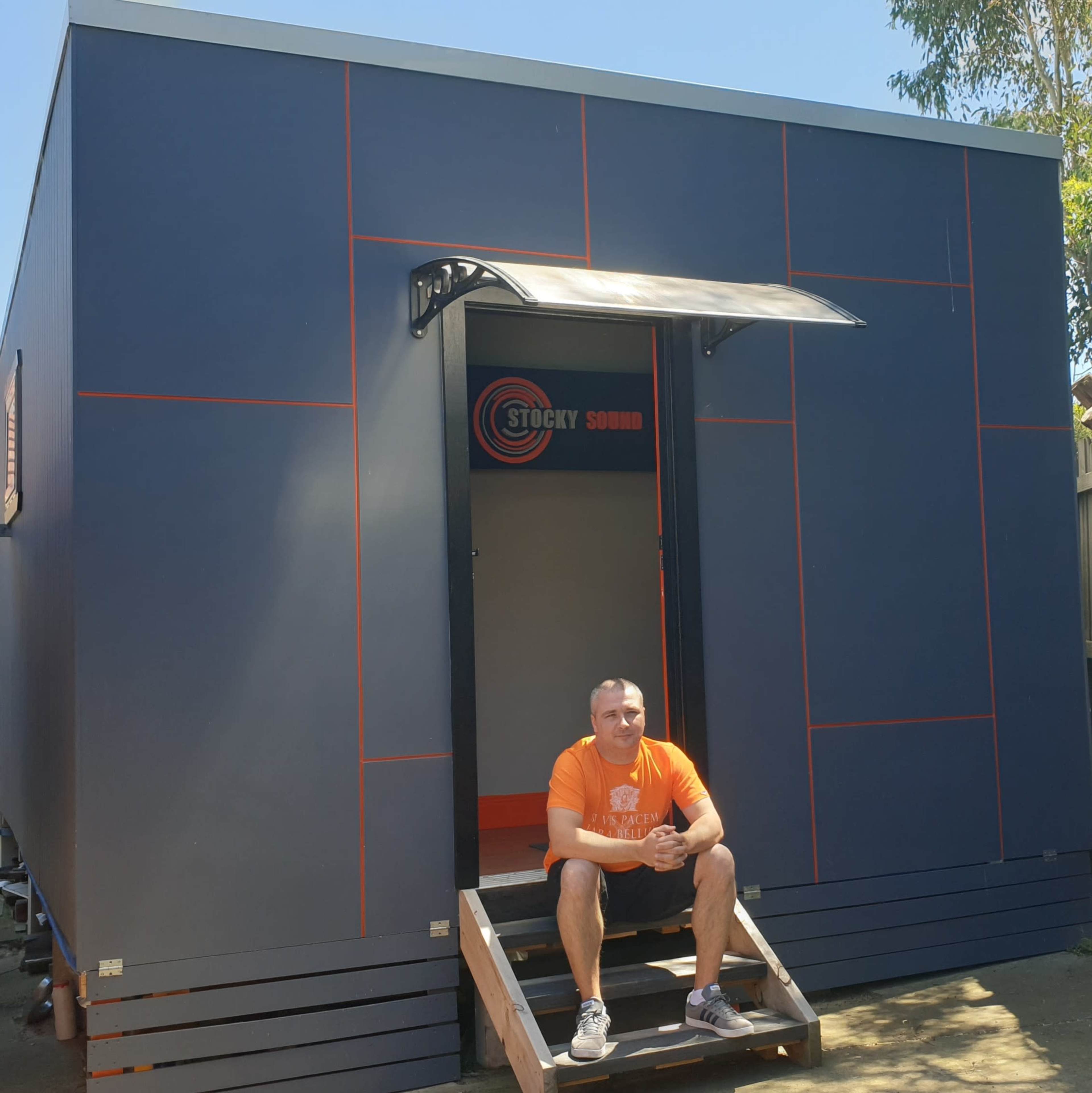 A man in an orange shirt sits on steps in front of a modern dark blue structure with a sign that reads "Stocky Sound."