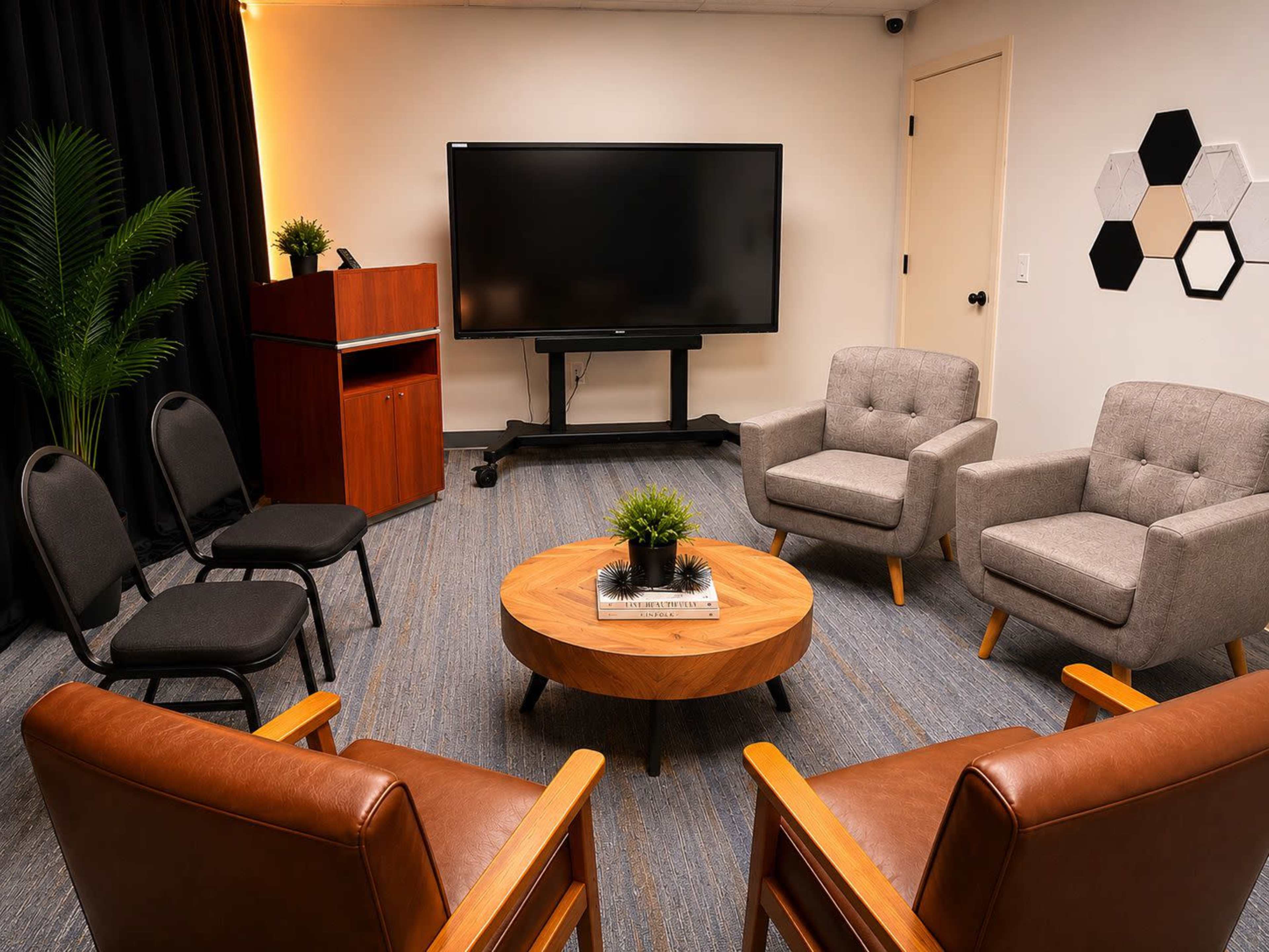 The image shows a meeting room with four chairs arranged around a circular wooden table, a large television on a stand, and a wooden cabinet against the wall.