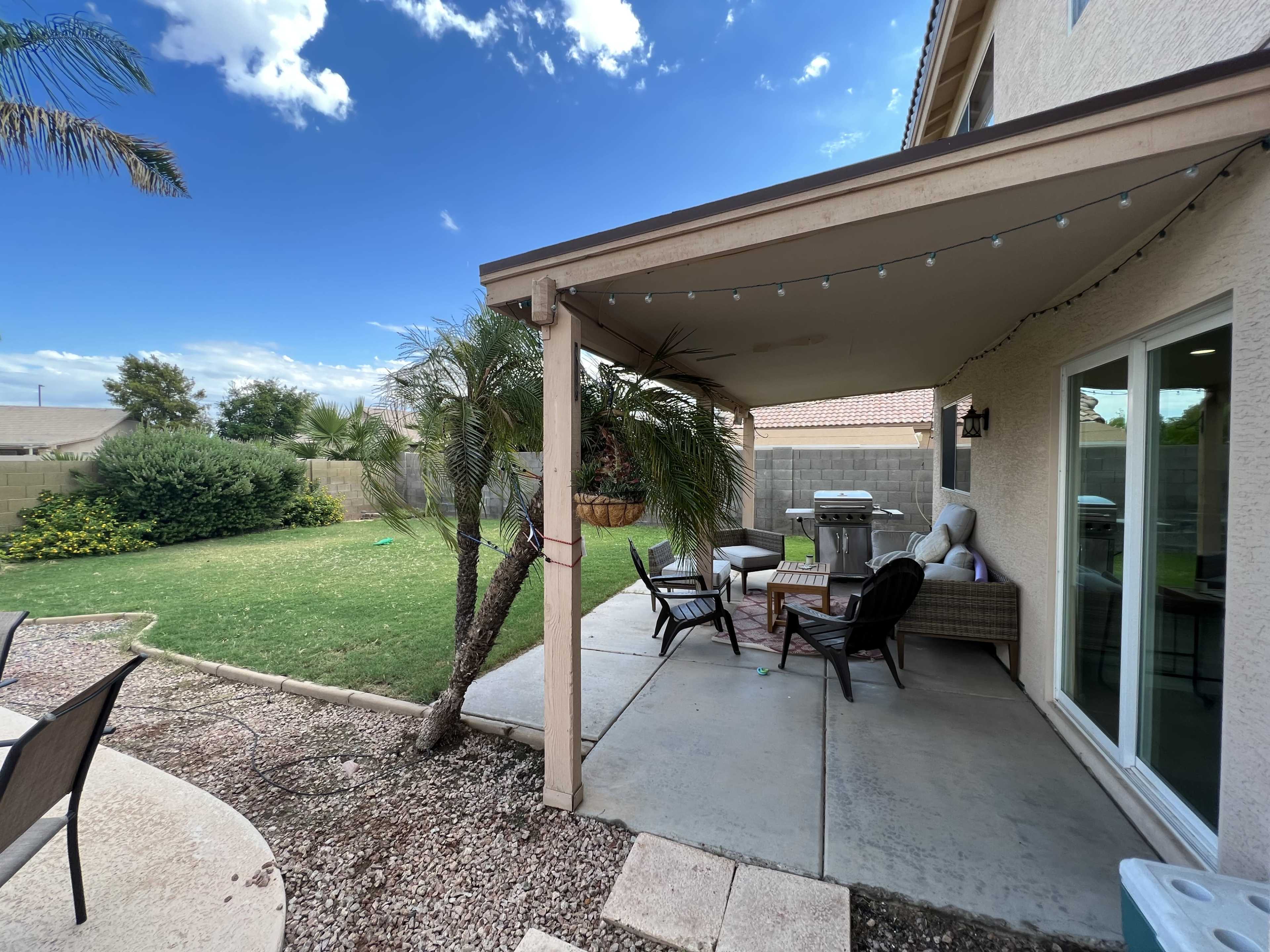 A patio area is shaded by a roof and features outdoor furniture, with a lawn and shrubs visible in the background.