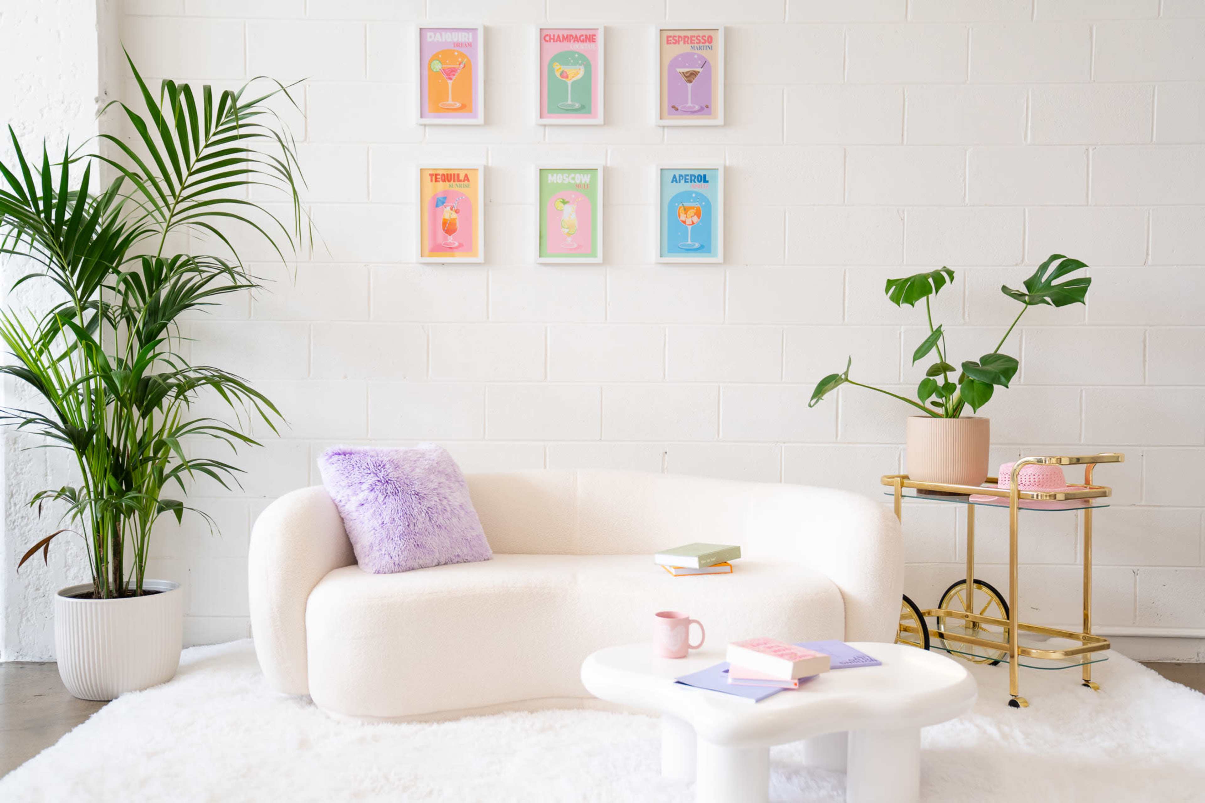 A modern seating area features a white sculptural couch, a round coffee table, and a side table with a pink telephone, accompanied by potted plants and colorful art prints on the wall.