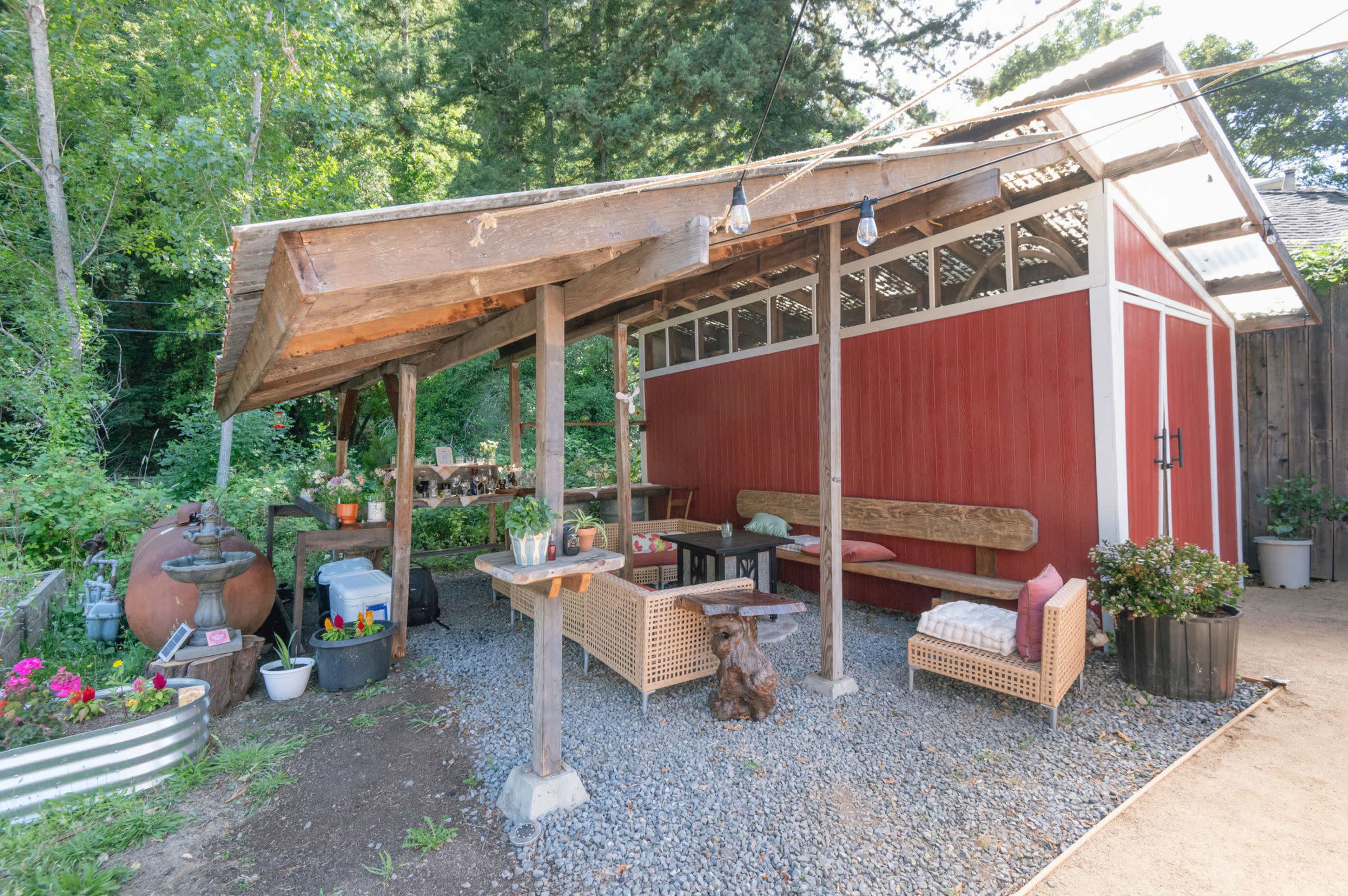 The image shows a rustic outdoor seating area next to a red wooden structure, featuring a sloped roof, wooden tables, and decorative plants.
