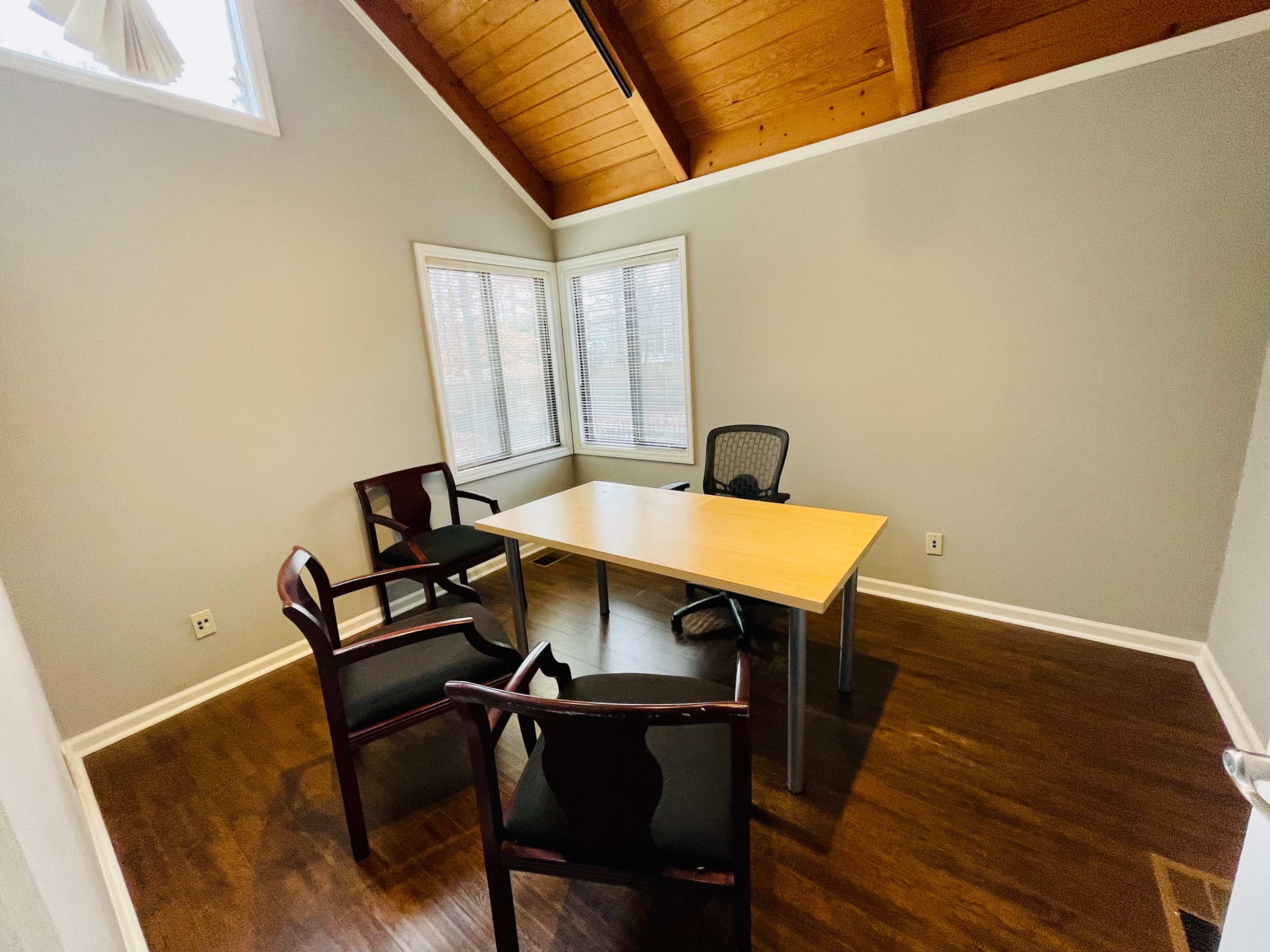 The image shows a small meeting room featuring a rectangular table surrounded by four chairs and large windows with blinds.