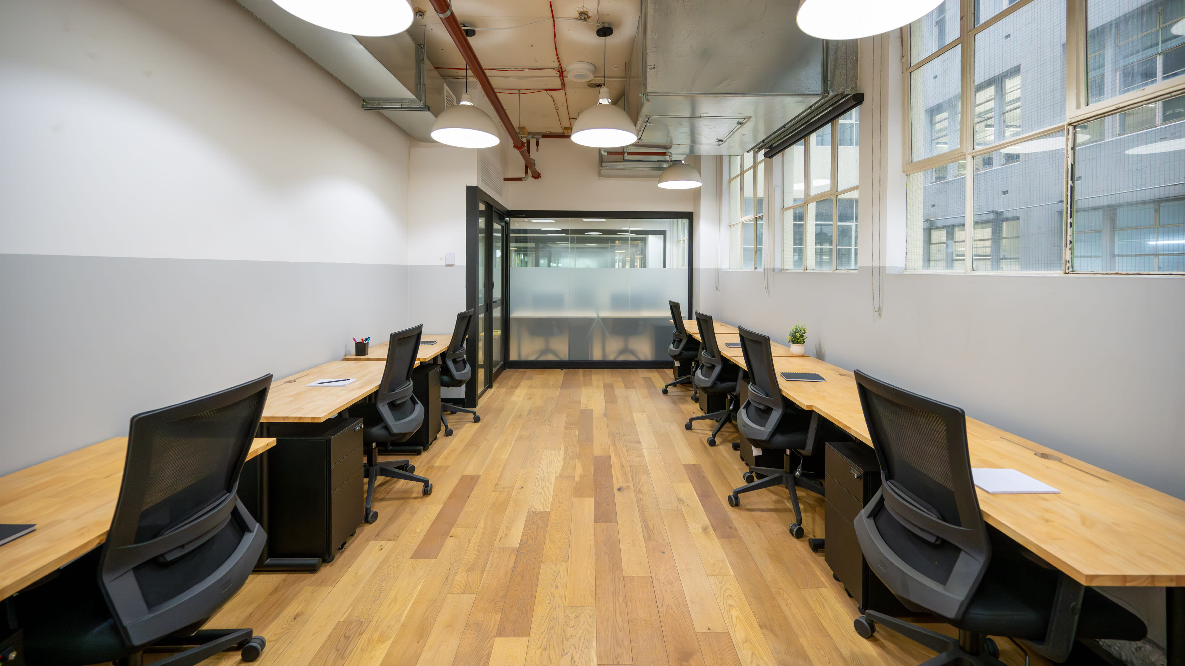 The image shows a modern office space with multiple wooden desks and ergonomic chairs arranged along a well-lit room featuring large windows.