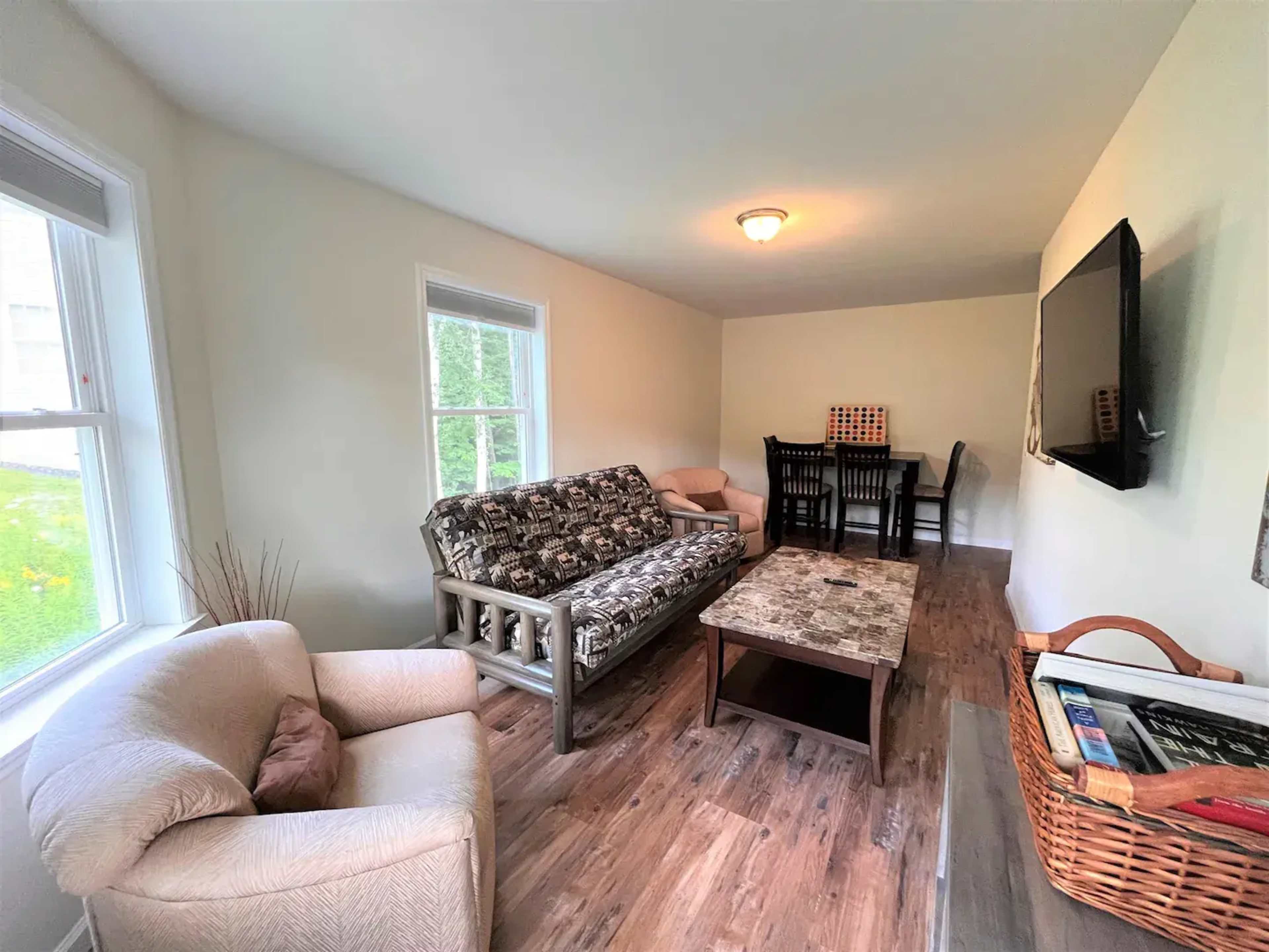A living room with a patterned sofa, a light-colored armchair, a wooden coffee table, and a dining table set for four.