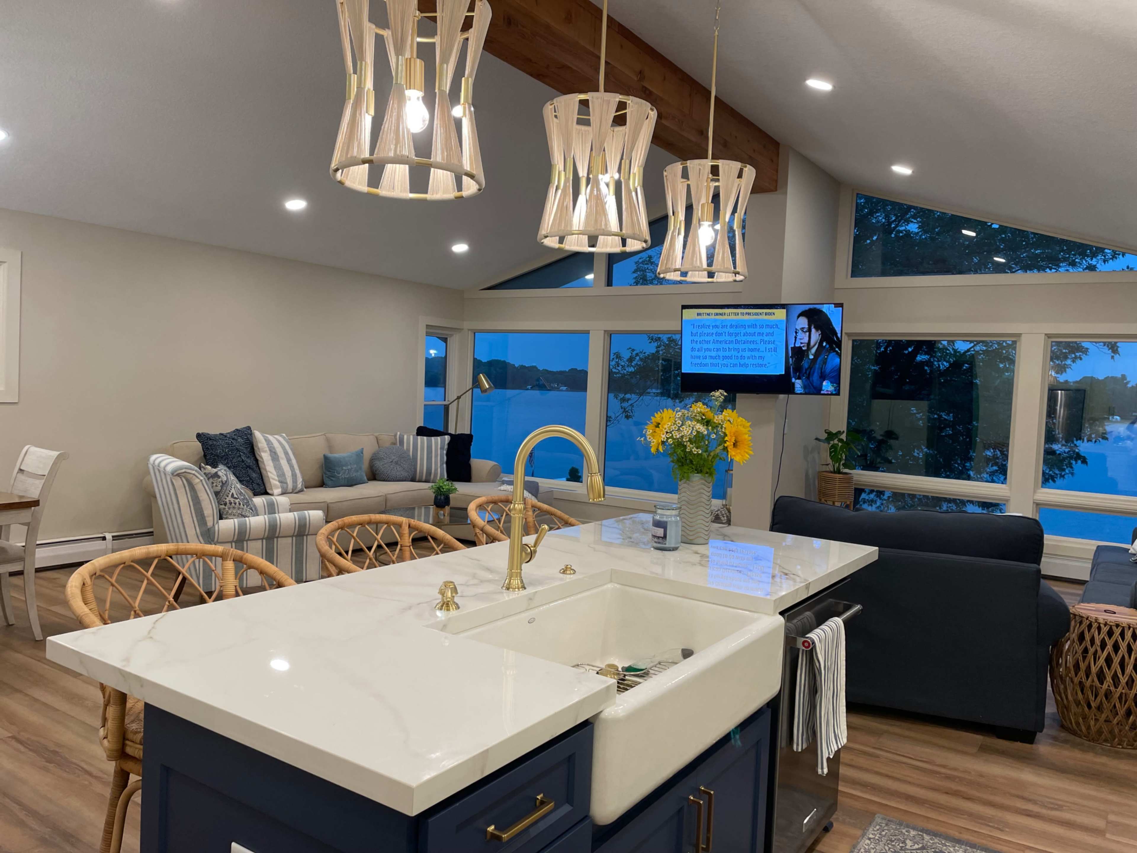 A modern kitchen with a marble countertop, a gold faucet, and a view of a living area featuring a sectional sofa and large windows overlooking a body of water.