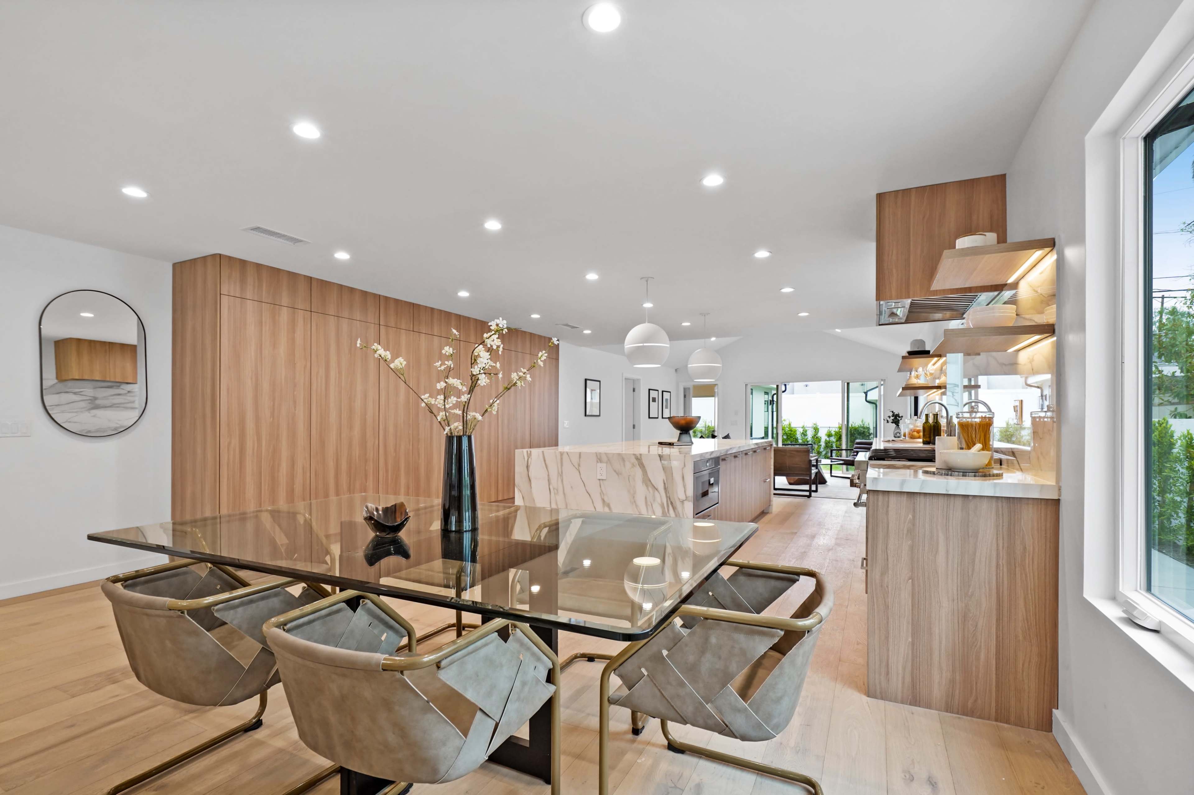 A modern kitchen and dining area features a glass dining table surrounded by upholstered chairs and light wood cabinetry.