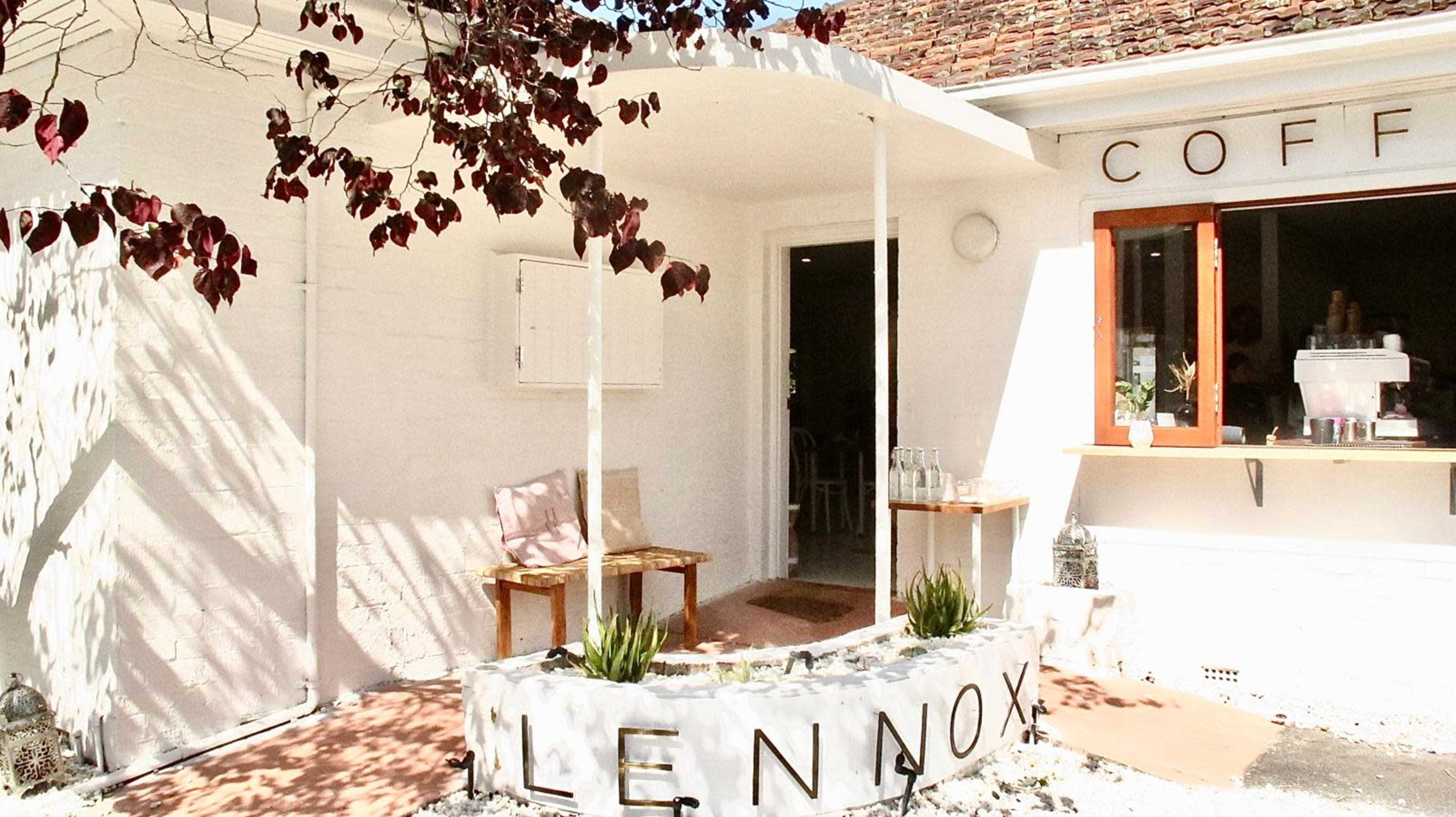 A modern café entrance with a curved stone bench and wooden window frame, surrounded by light-colored walls and foliage.
