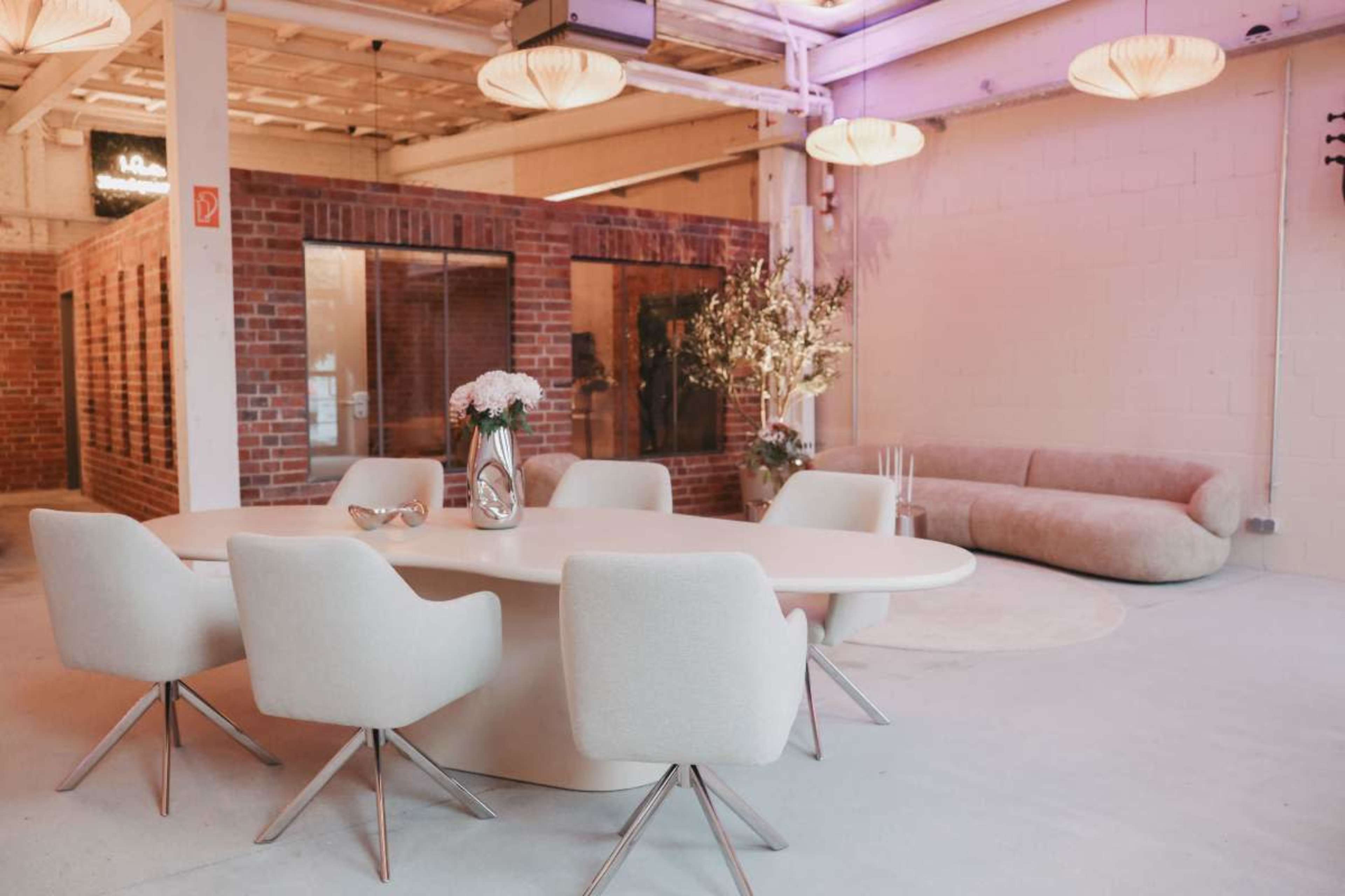 A spacious, modern meeting room features a large oval table surrounded by six light-colored chairs, with a plush sofa and brick walls in the background.