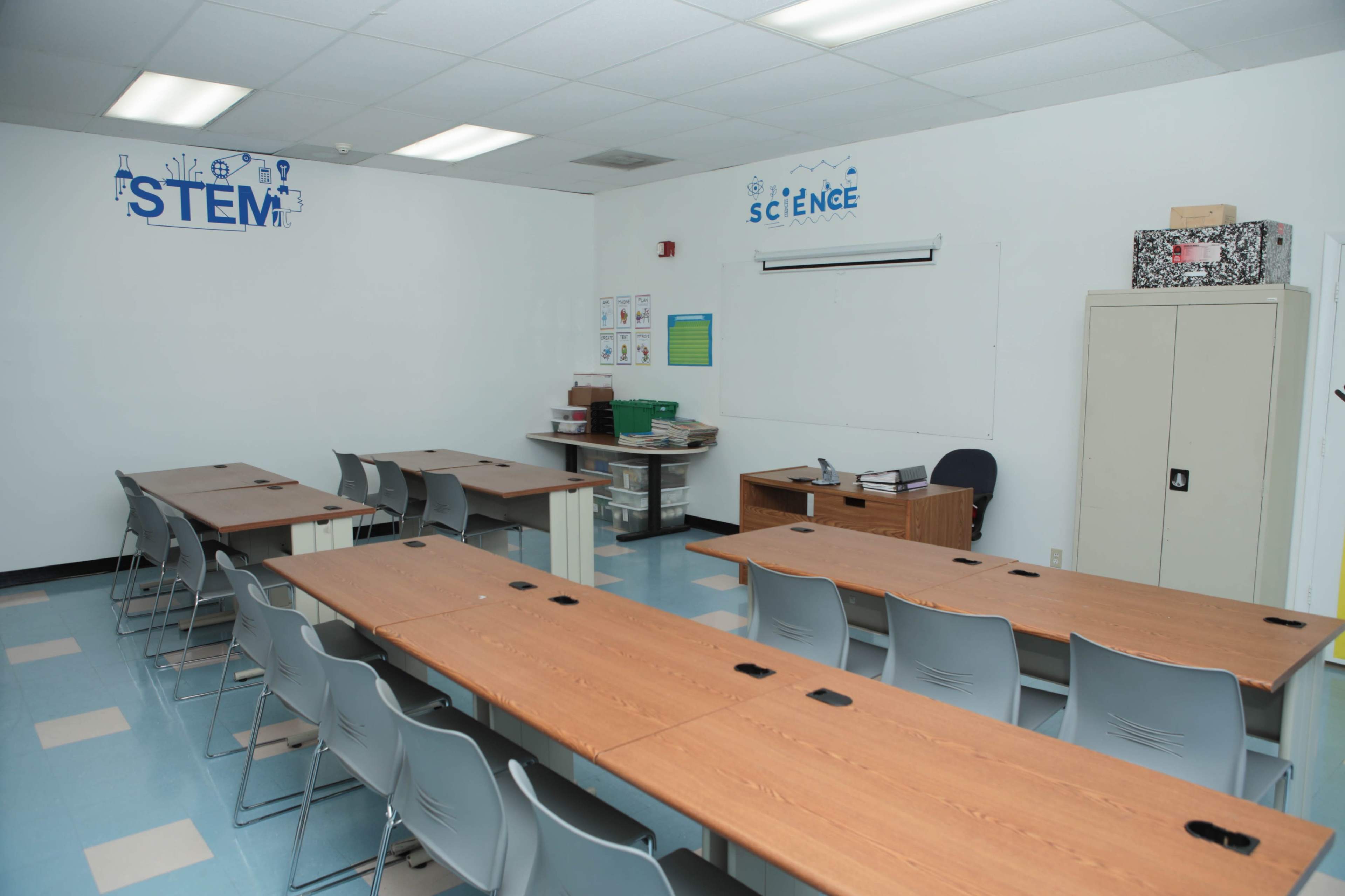 A classroom features rows of tables and chairs, a teacher's desk at the front, and educational posters labeled "STEM" and "Science" on the walls.