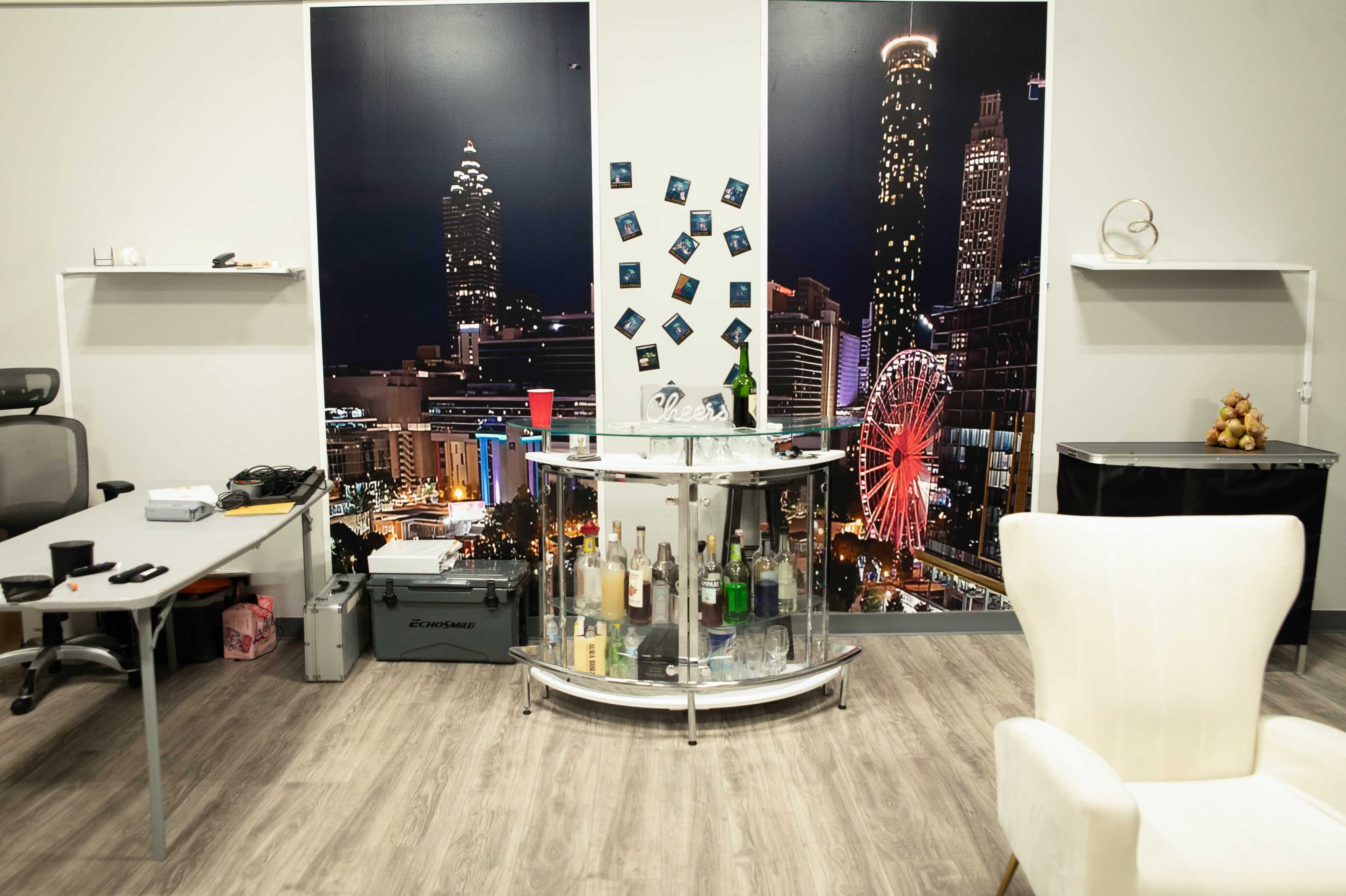 The image shows an office space featuring a circular bar cart in front of a large mural depicting a city skyline at night, along with a white chair and various office supplies on a nearby table.