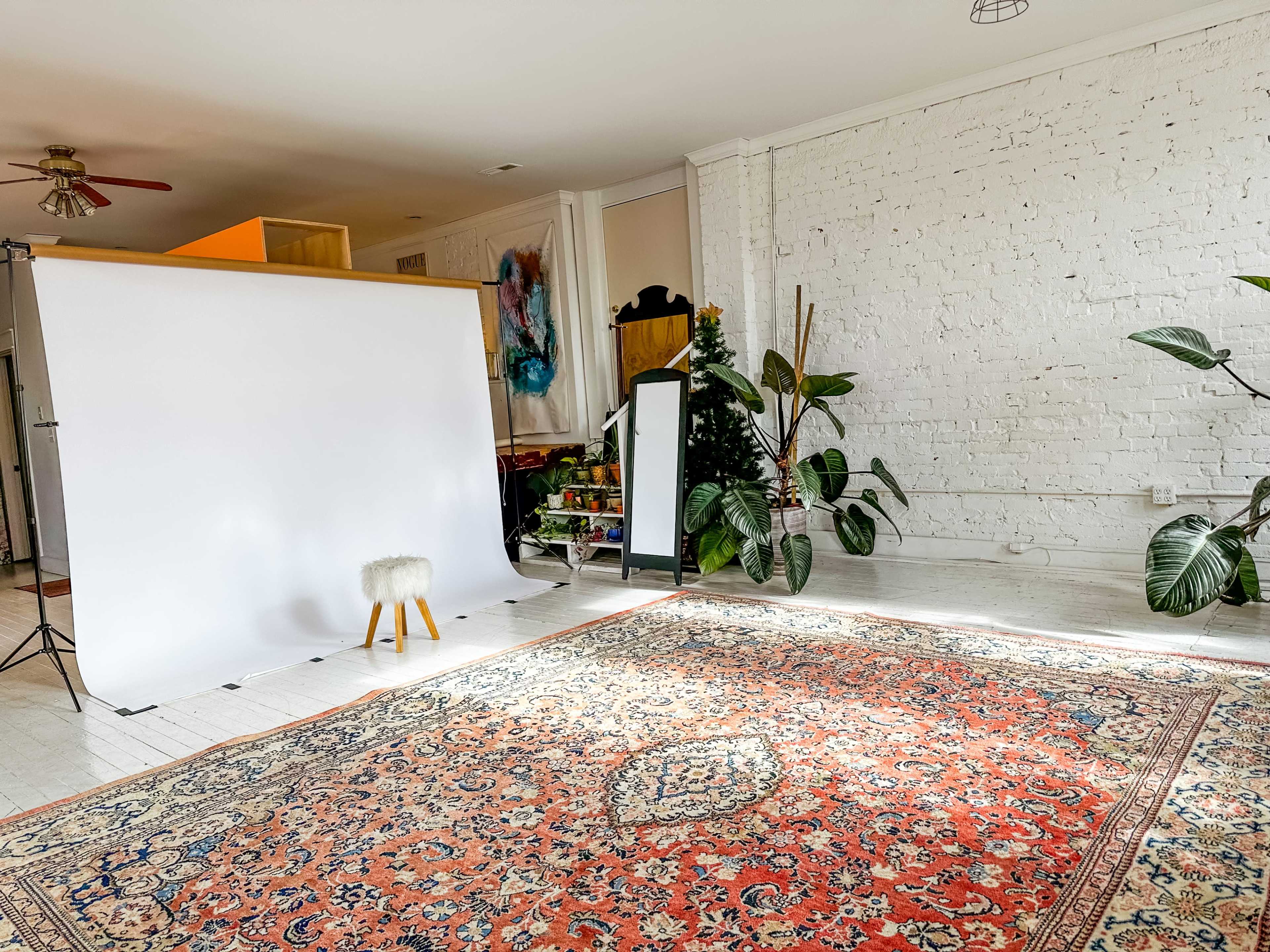 A spacious room with a white backdrop, an ornate rug, a small stool, a floor mirror, and large potted plants against white brick walls.
