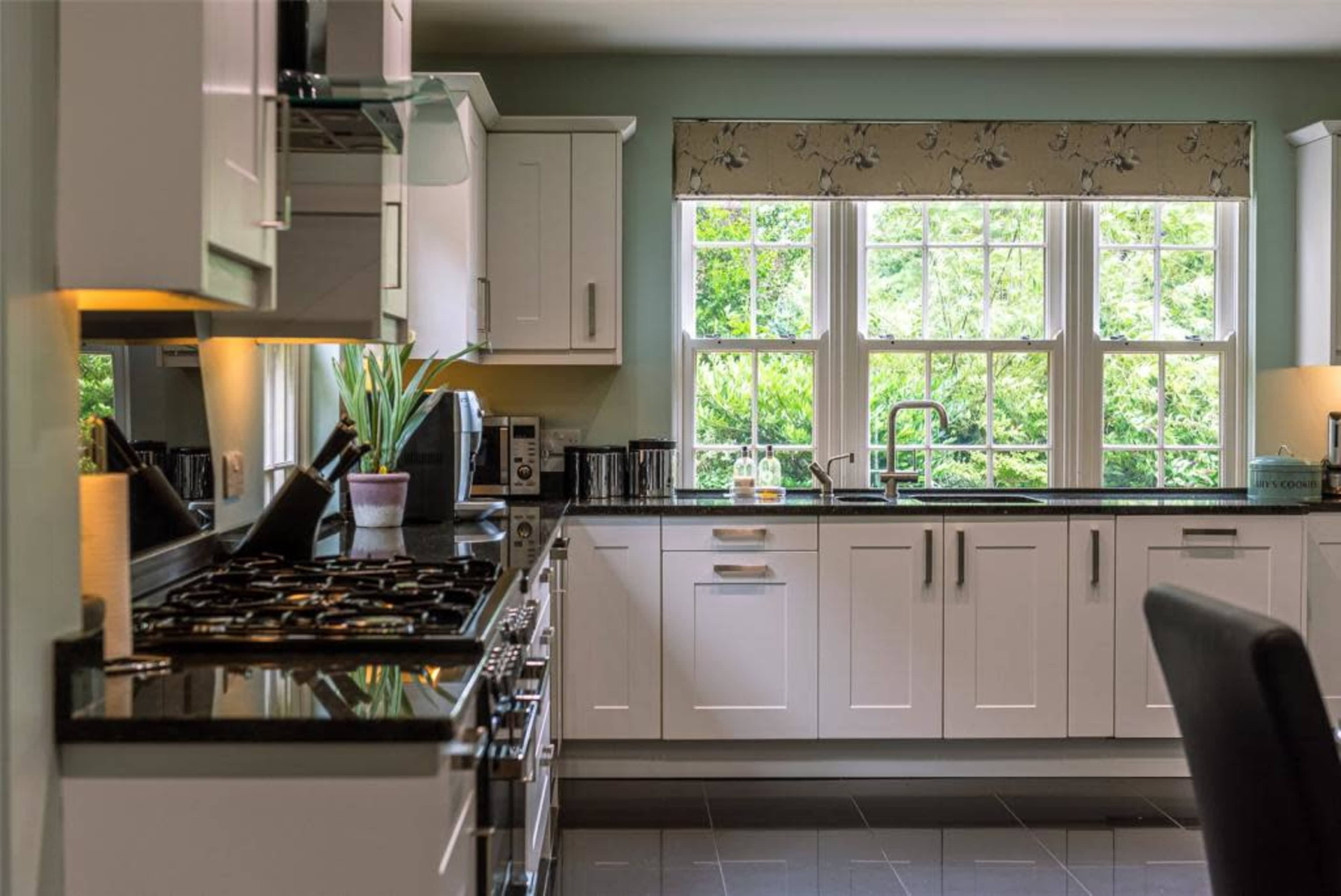 A modern kitchen features white cabinetry, a black countertop, and large windows overlooking greenery outside.