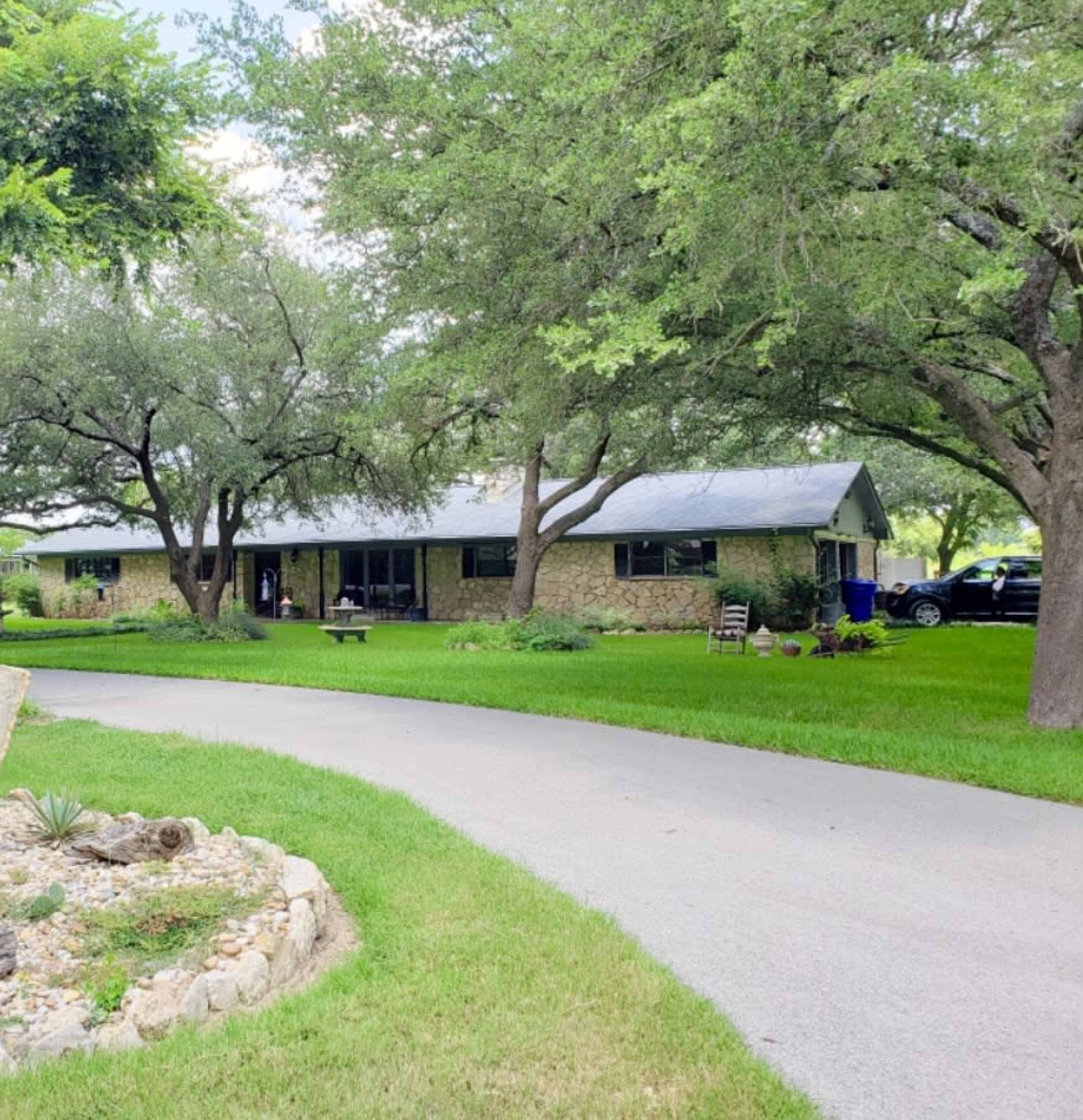A single-story stone house is set back from a curved driveway surrounded by green grass and trees.