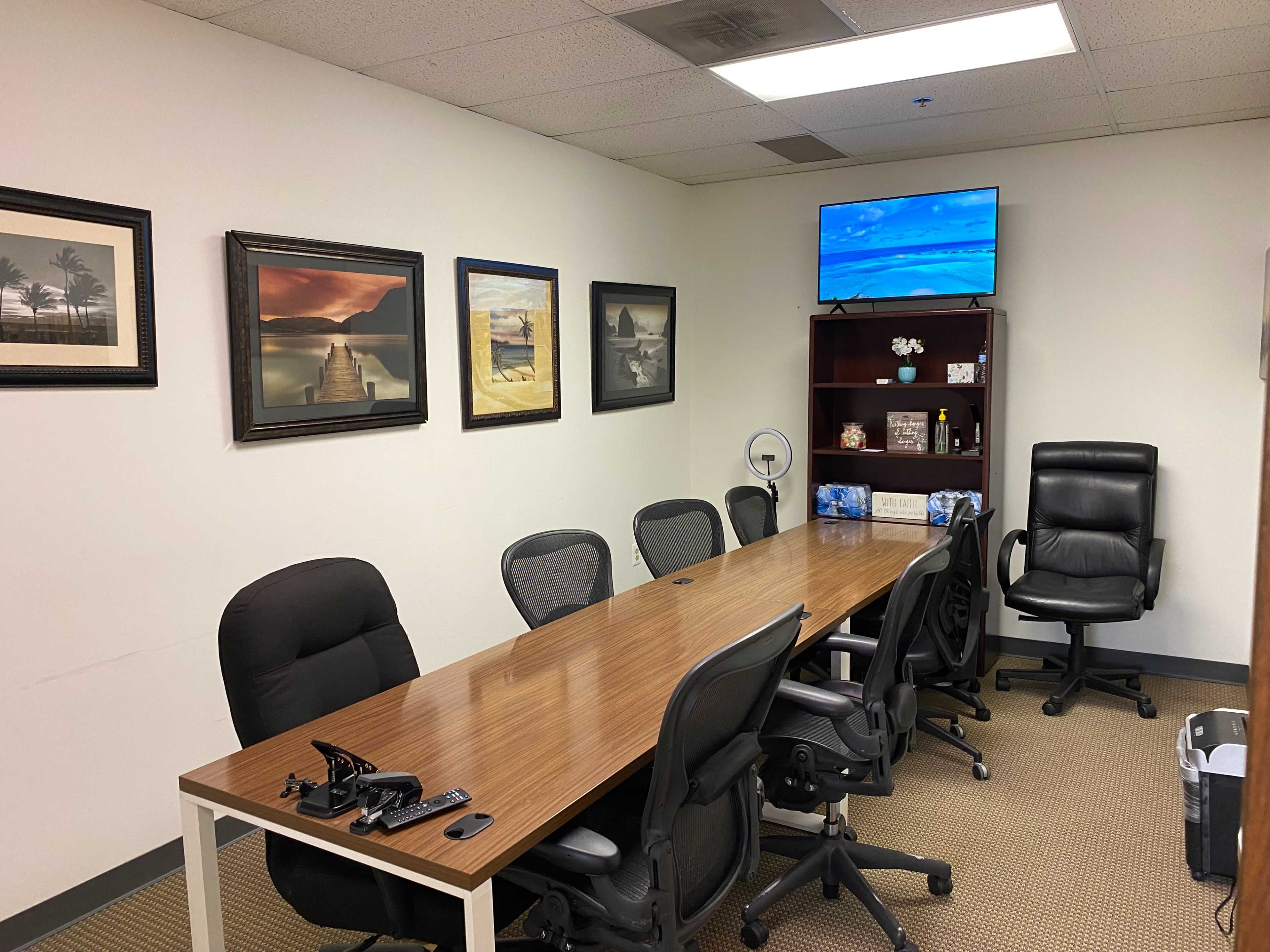 The image shows a conference room with a long wooden table, several office chairs, framed pictures on the walls, and a television mounted in one corner.