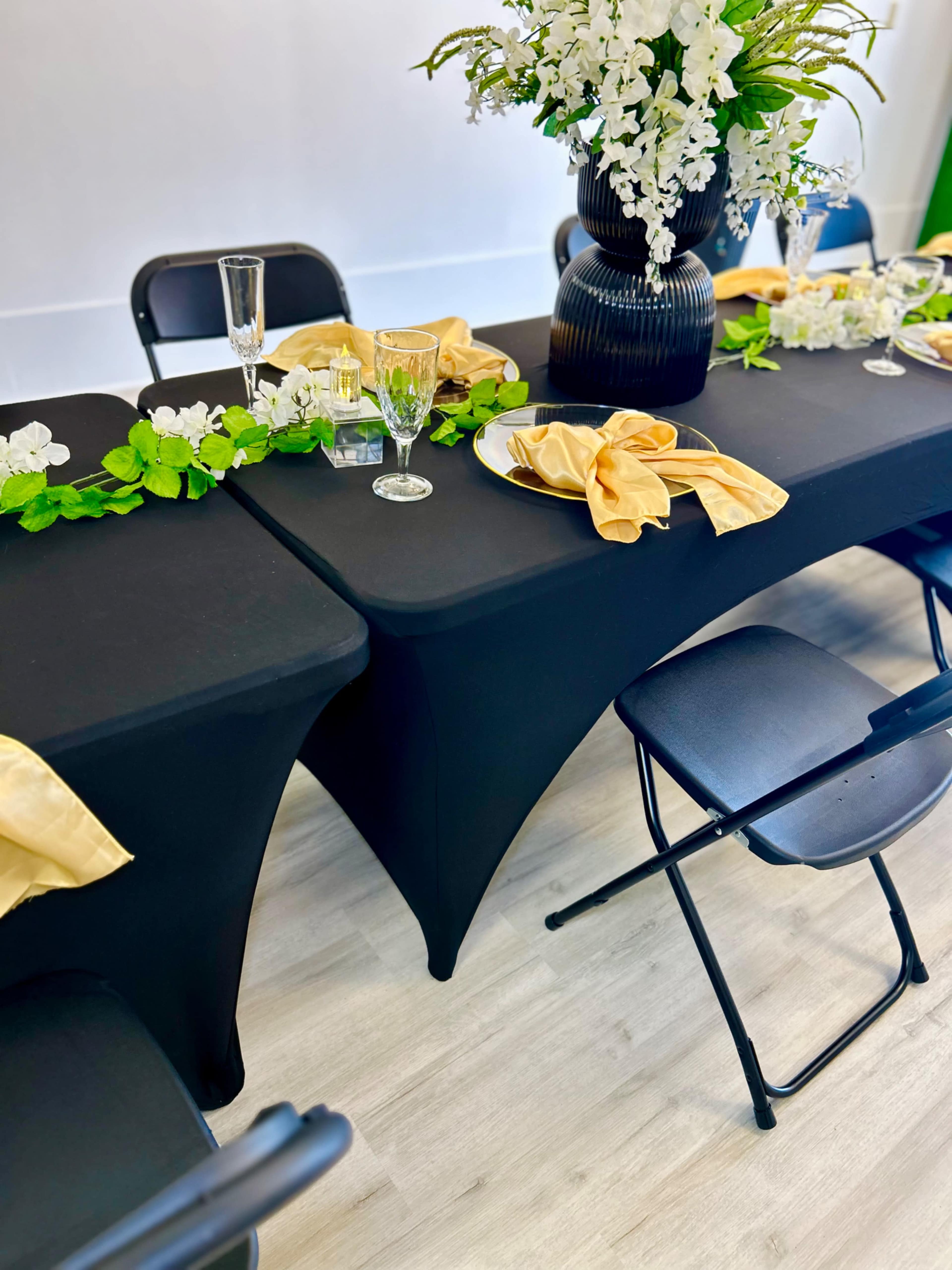 A black table is set with yellow napkins and floral decorations, accompanied by chairs, glassware, and centerpieces of white flowers.