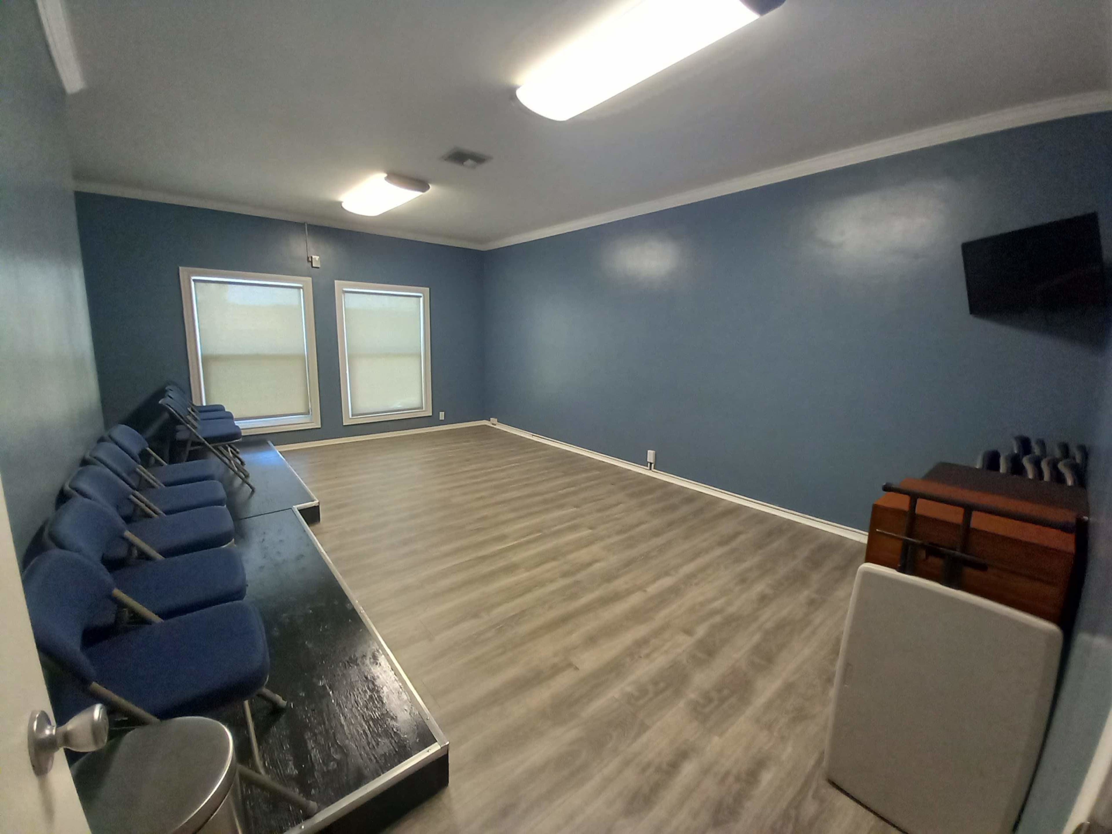 A sparsely furnished waiting room with blue walls, several blue chairs arranged in a row, and windows letting in natural light.