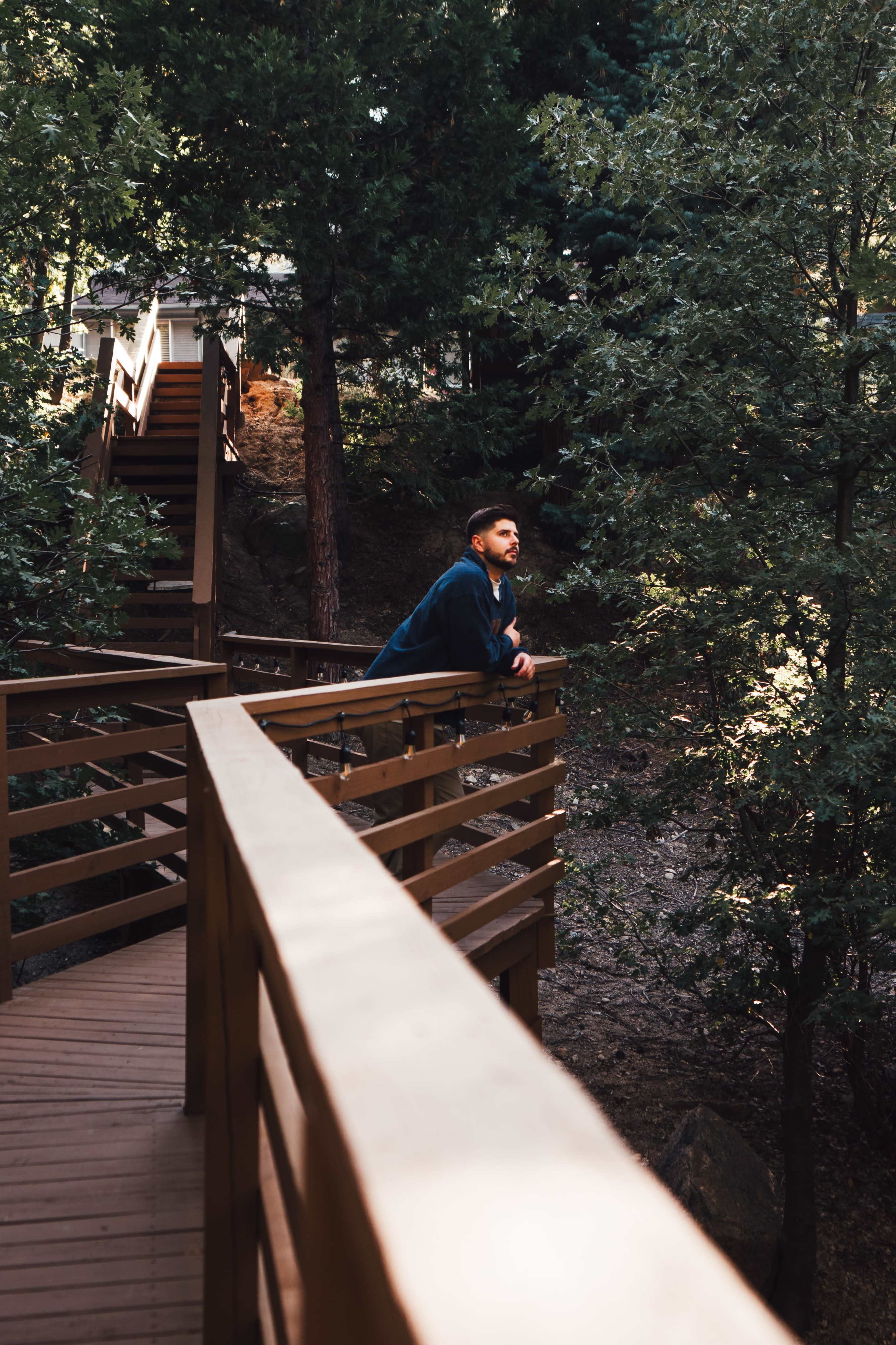 A man leans on a wooden railing overlooking a forested area with stairs visible in the background.