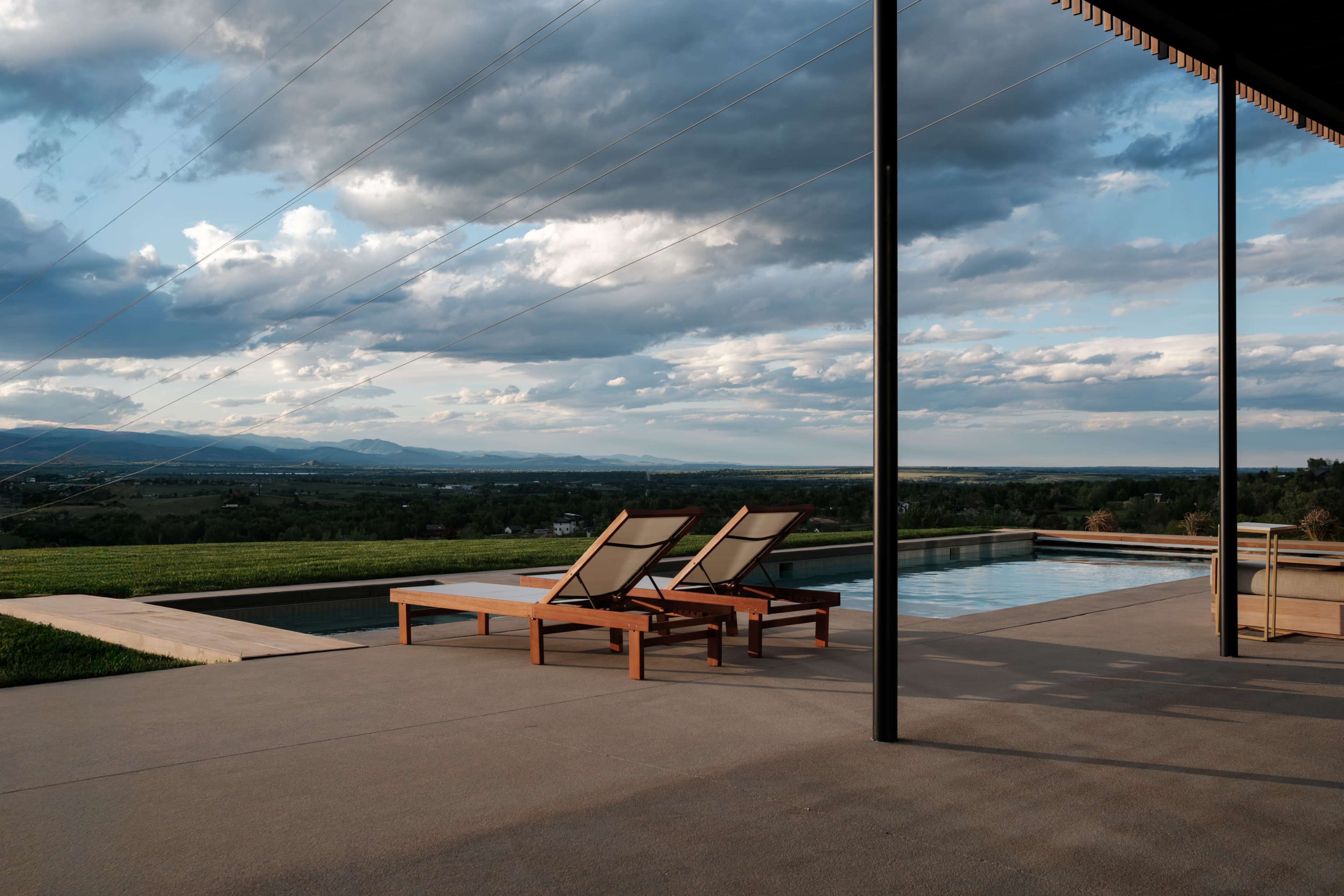 A minimalist patio features two lounge chairs beside a swimming pool overlooking a scenic landscape with rolling hills and cloudy skies.