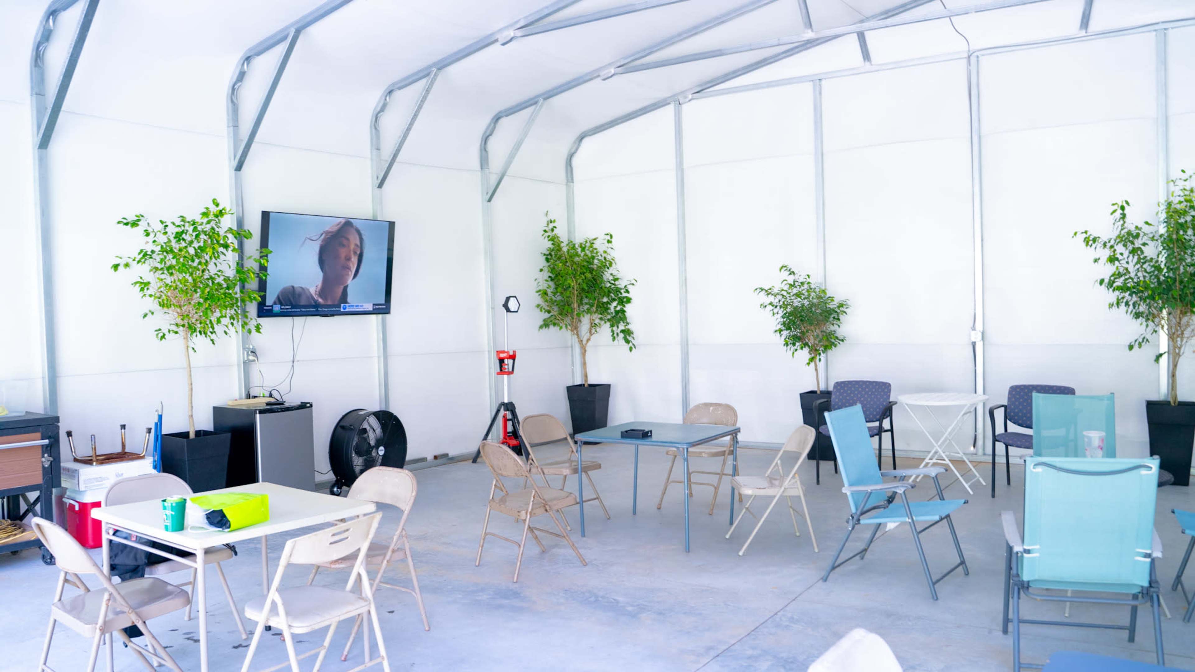 The interior of a large, open shed features several tables and chairs, a television mounted on the wall, and indoor plants positioned around the space.