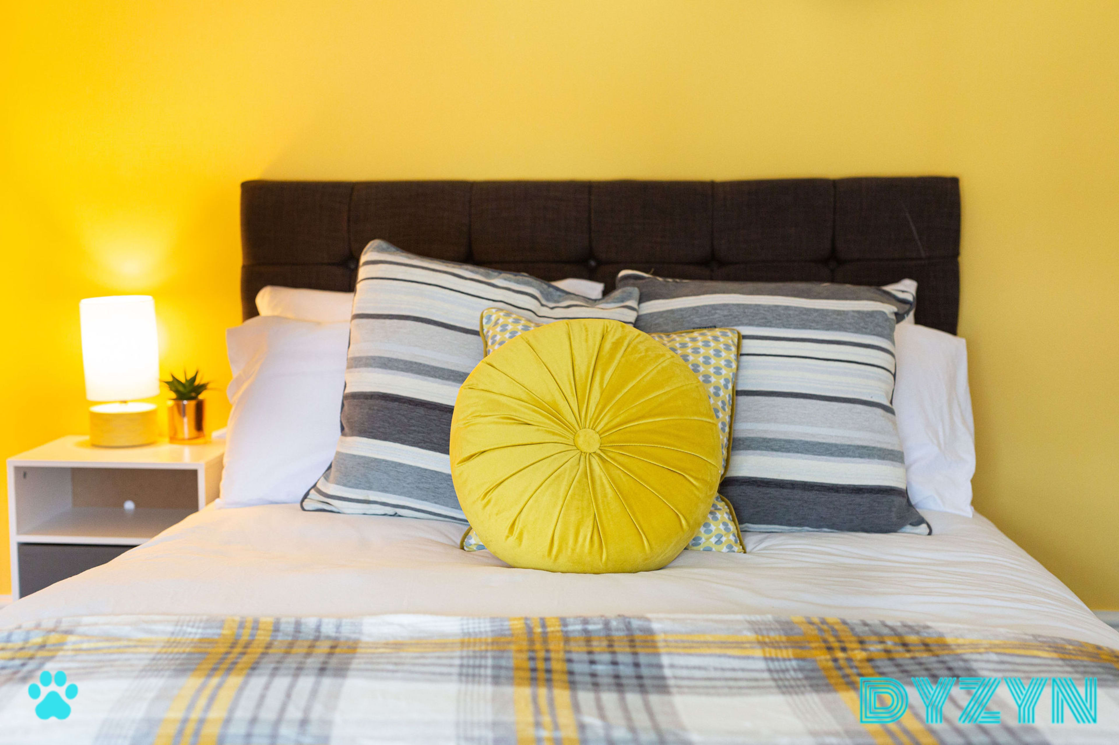 A neatly made bed with a dark headboard, striped pillows, and a large yellow round cushion against a bright yellow wall.