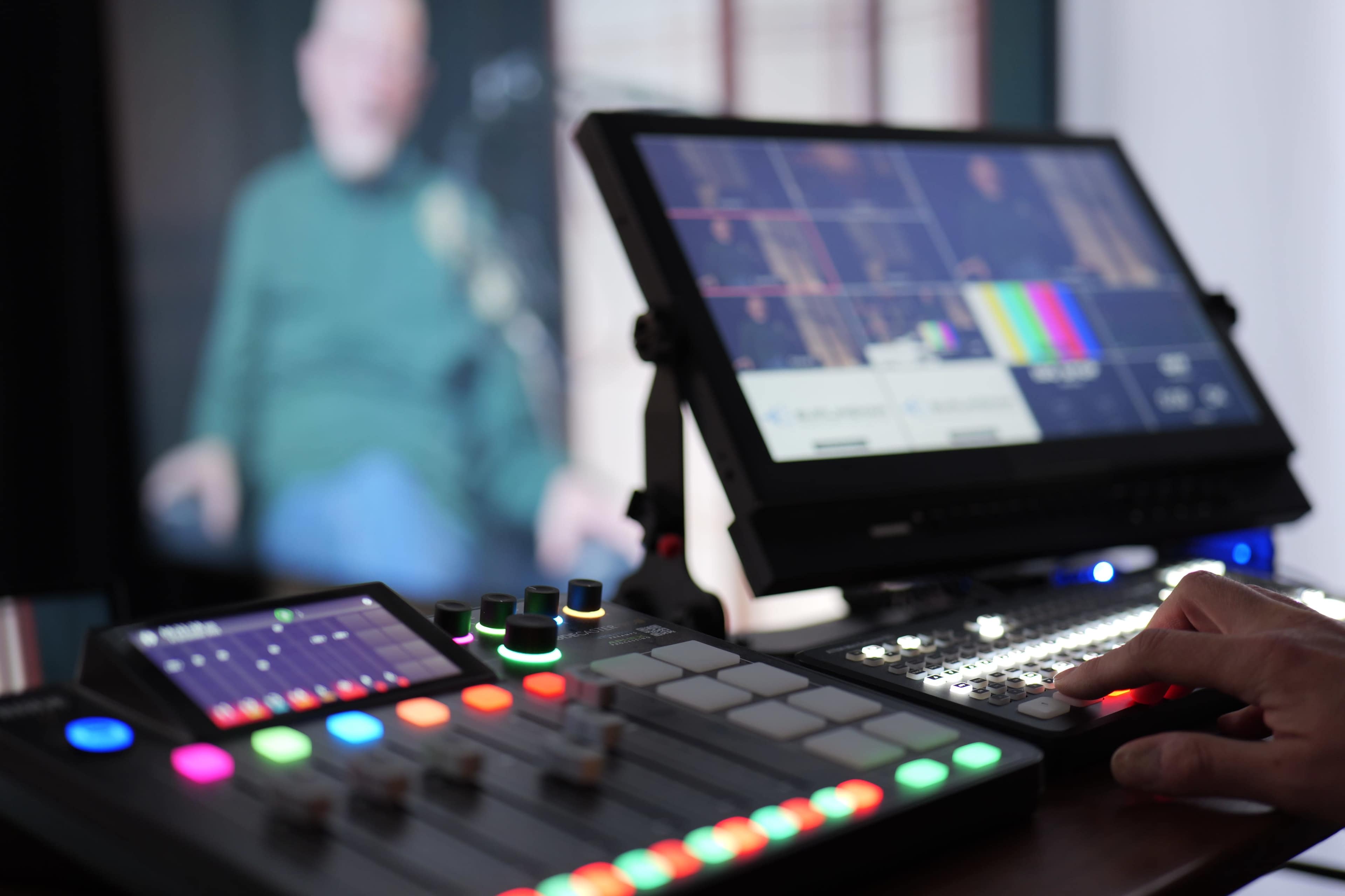 The image shows a close-up of a video production control panel with colorful buttons and screens, while a blurred figure sits in the background.