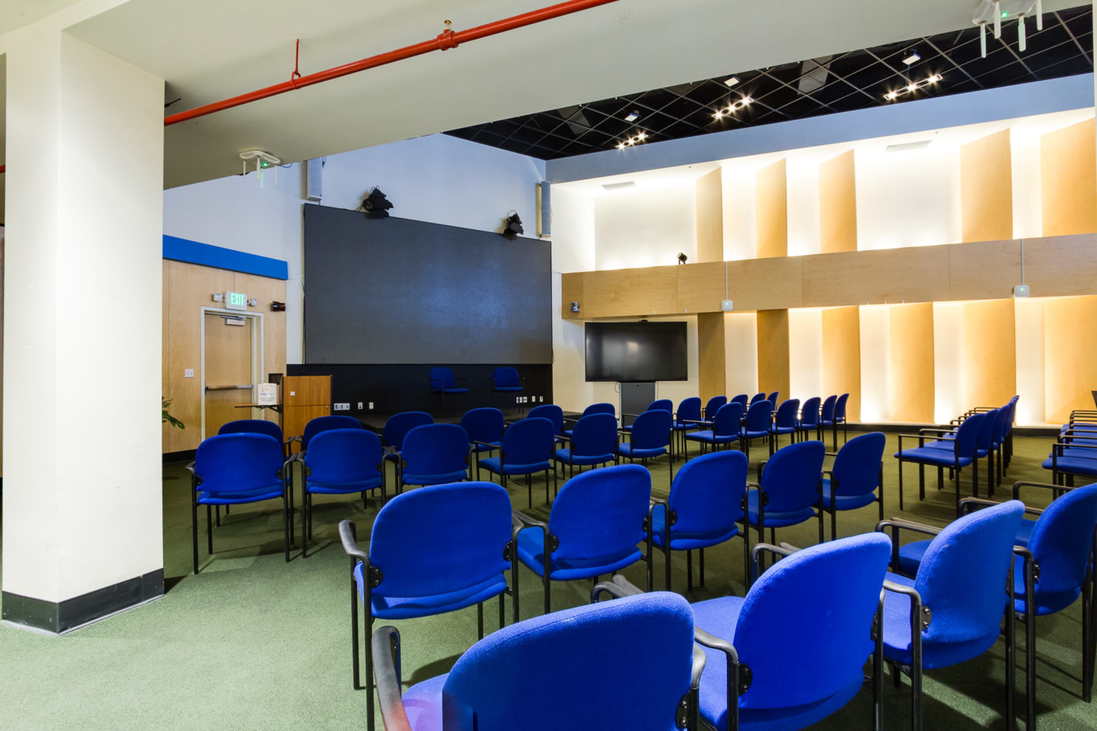 A modern seminar room with rows of blue chairs facing a central screen and a small stage in a well-lit space.