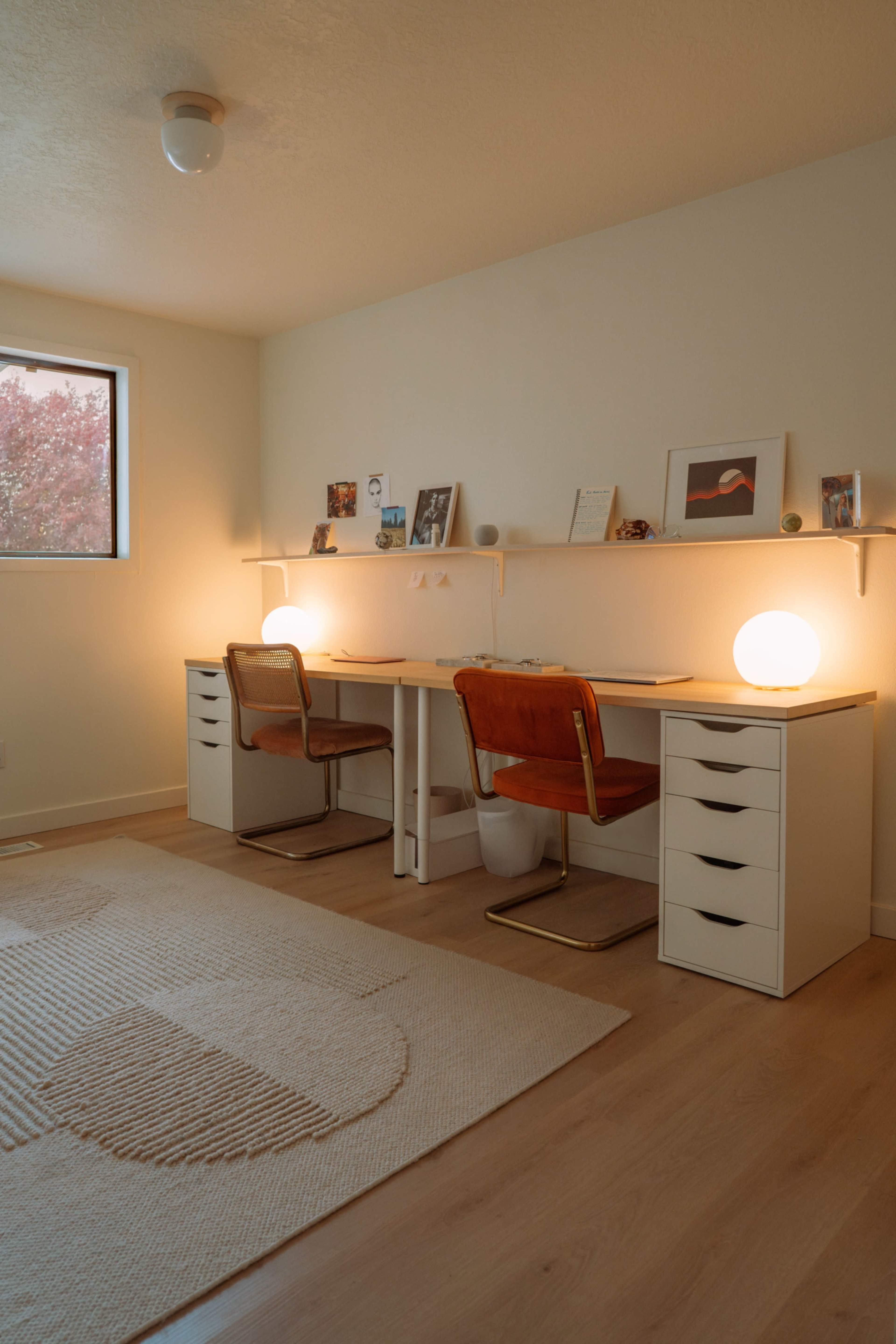 The image shows a tidy home office with two desks, each equipped with a chair and a lamp, set against a wall with framed pictures and décor.