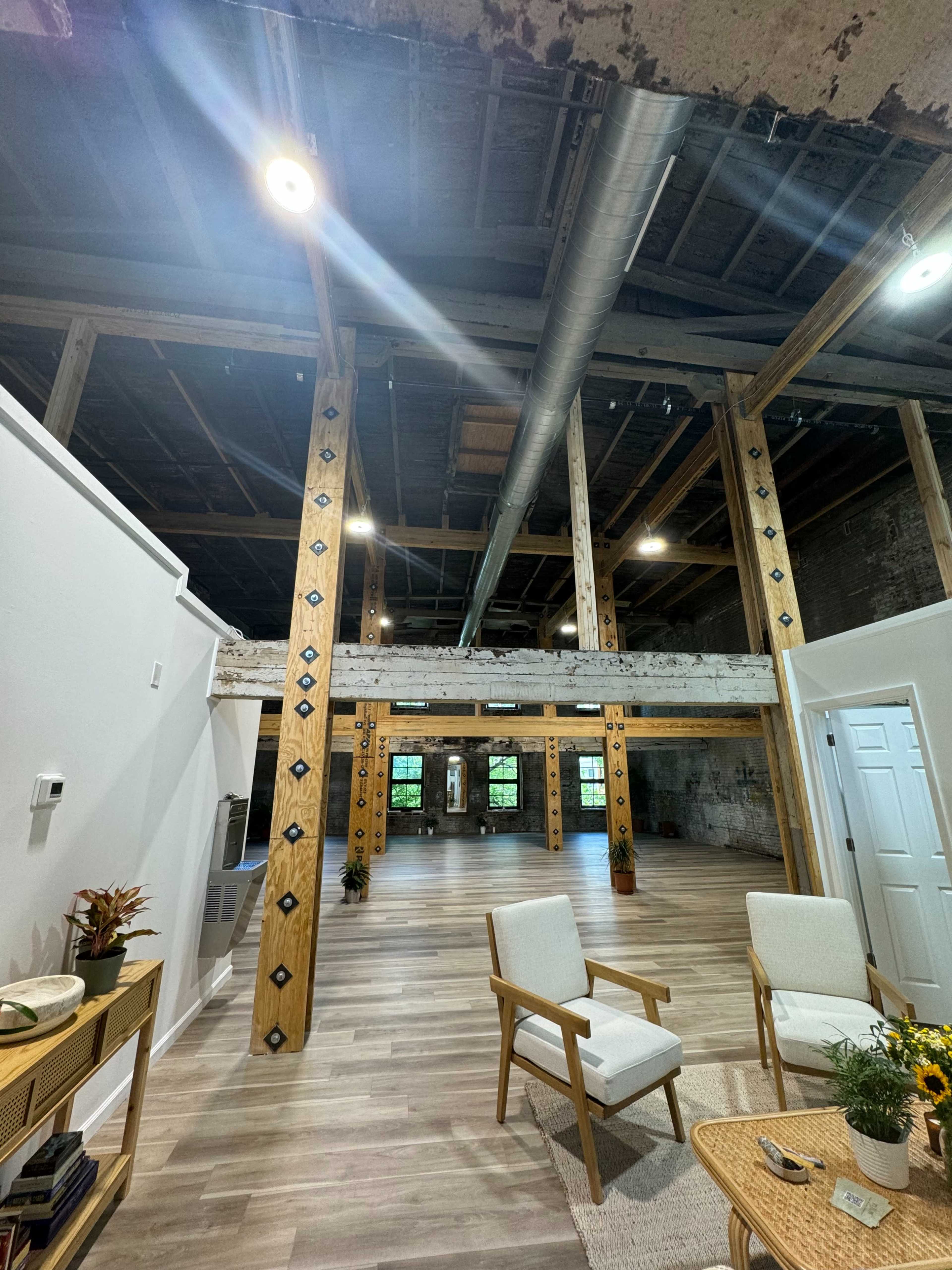 An interior space with wooden support beams, large windows, and minimal furniture, featuring a light-colored floor and exposed industrial elements.