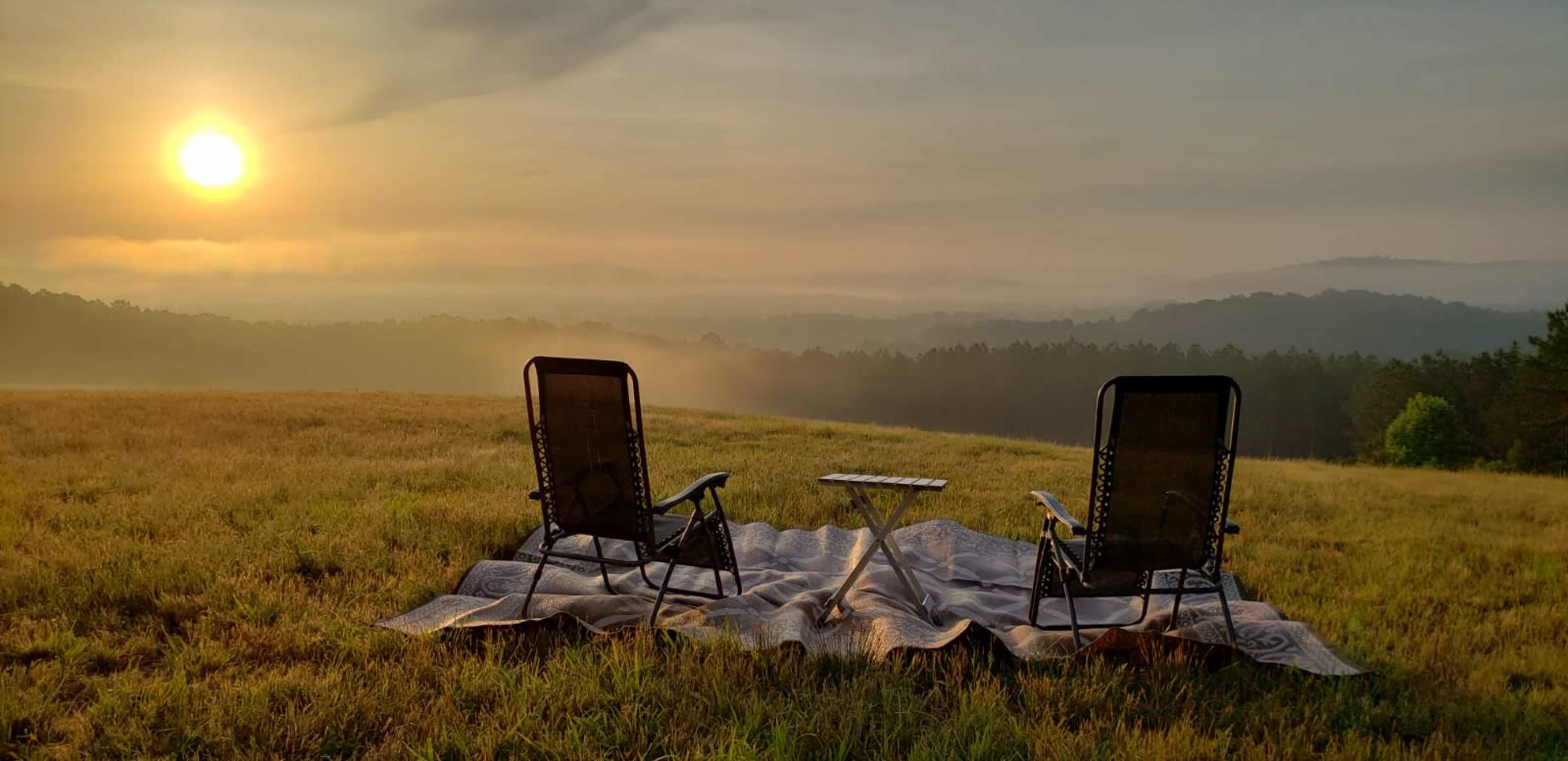 Two chairs are set up on a blanket in a grassy field, facing a sunrise over distant hills.