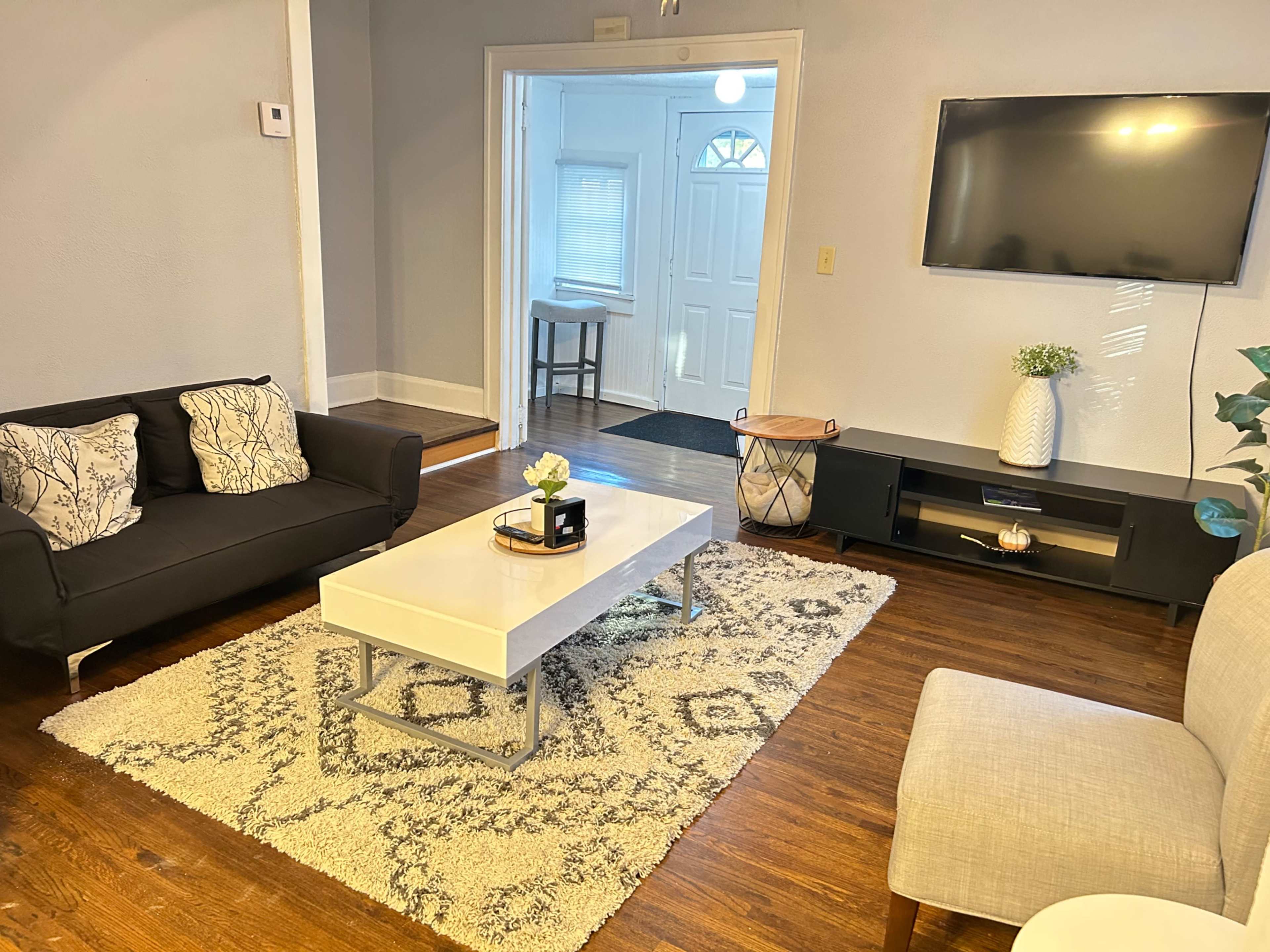 A living room with a black sofa, a white coffee table, a light gray armchair, and a TV mounted on the wall, all arranged on a patterned rug.
